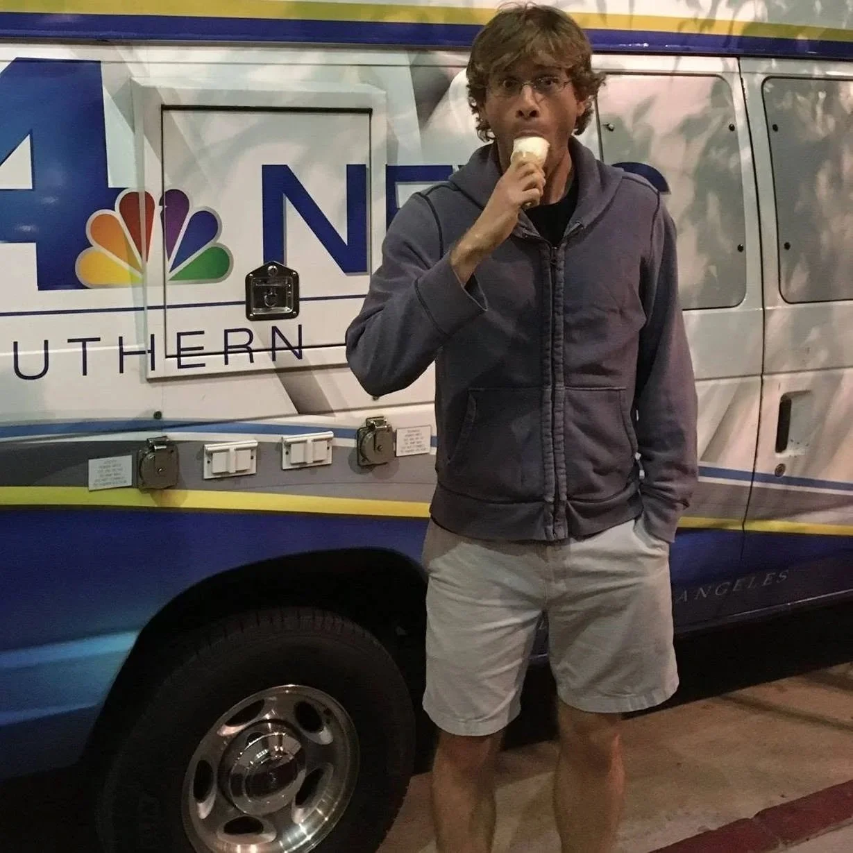 A man with glasses and curly hair wearing a gray jacket and beige shorts, standing in front of a NBC 4 Southern news van, eating an ice cream cone.