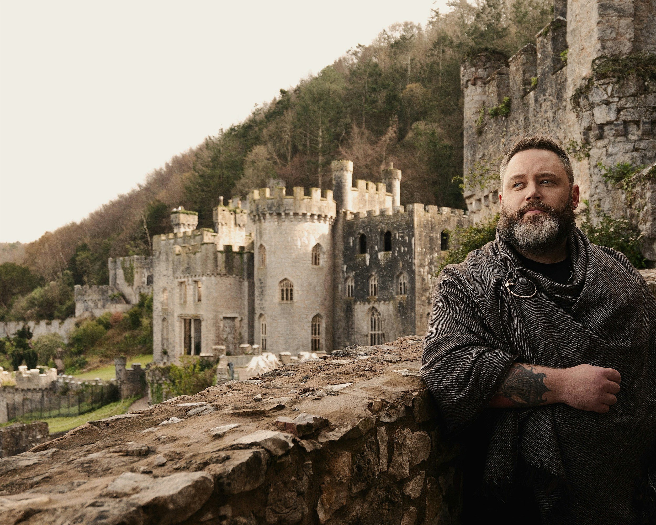 A bearded man with tattoos wearing a dark cloak standing next to a stone wall in front of a medieval castle on a hillside.