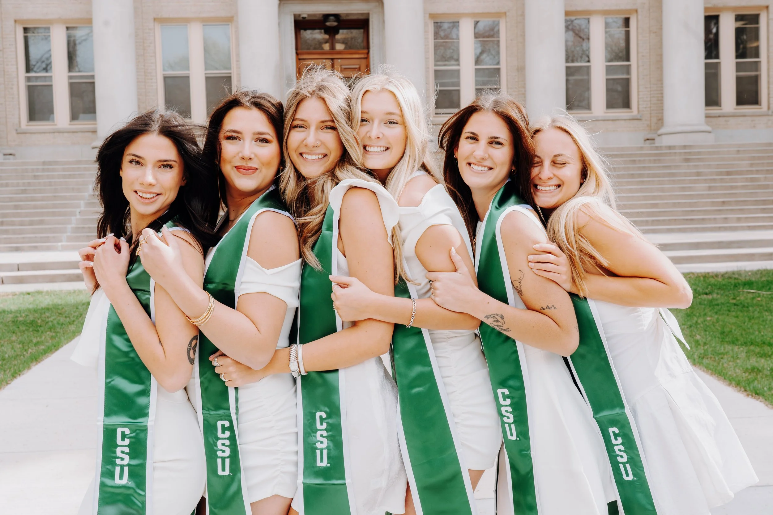 Group of six young women in white dresses with green CSU sashes standing on steps in front of a large building, smiling and embracing each other.