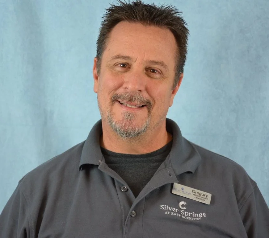Man smiling with goatee, wearing a gray polo shirt with "Silver Springs at East Norriton" logo and a name tag, against a light blue background.
