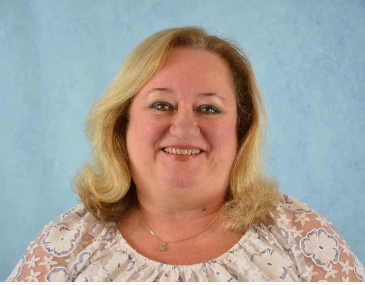 Smiling person with shoulder-length blonde hair wearing a floral blouse, posing against a blue background.