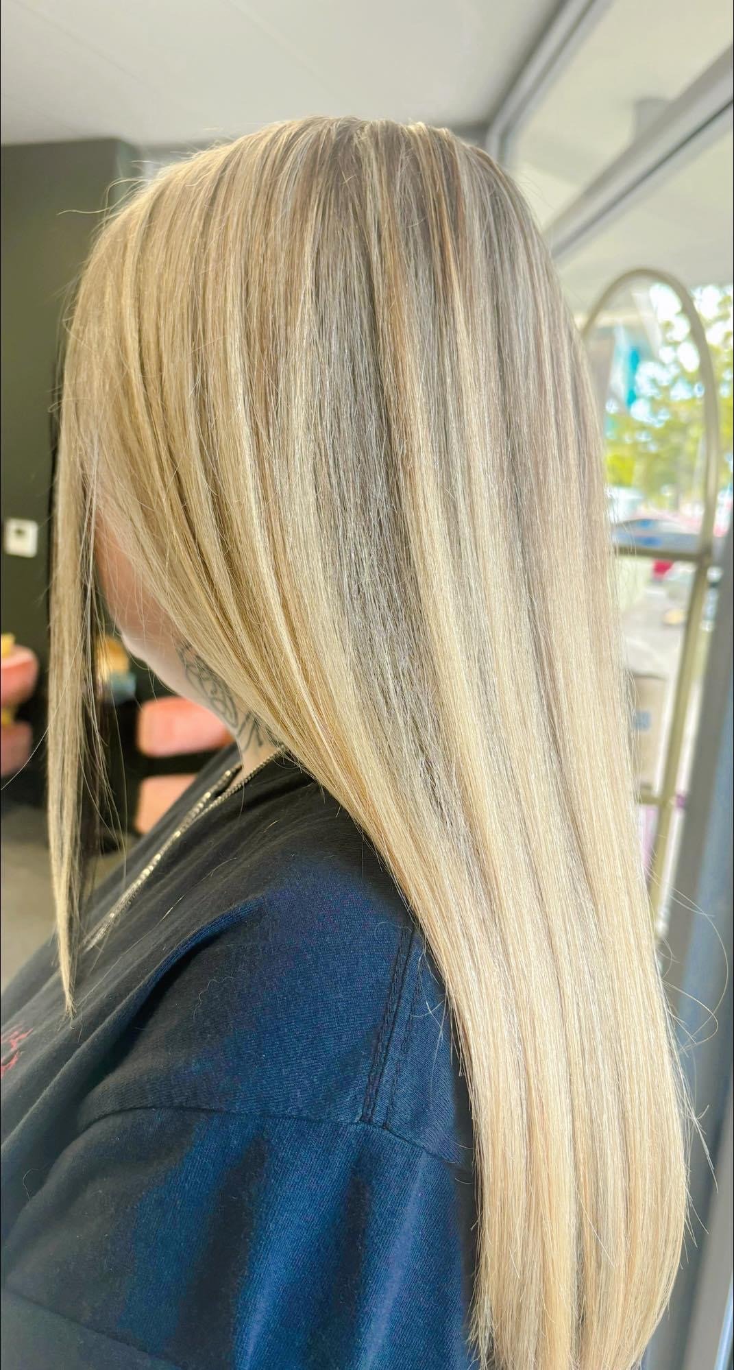 Close-up of a person with long, straight, blonde hair and a tattoo on their neck, wearing a dark-colored top.