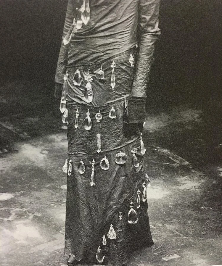 Close-up of a person's lower body dressed in a textured, flowing garment decorated with hanging jewelry and pendants, standing on a surface with reflections.