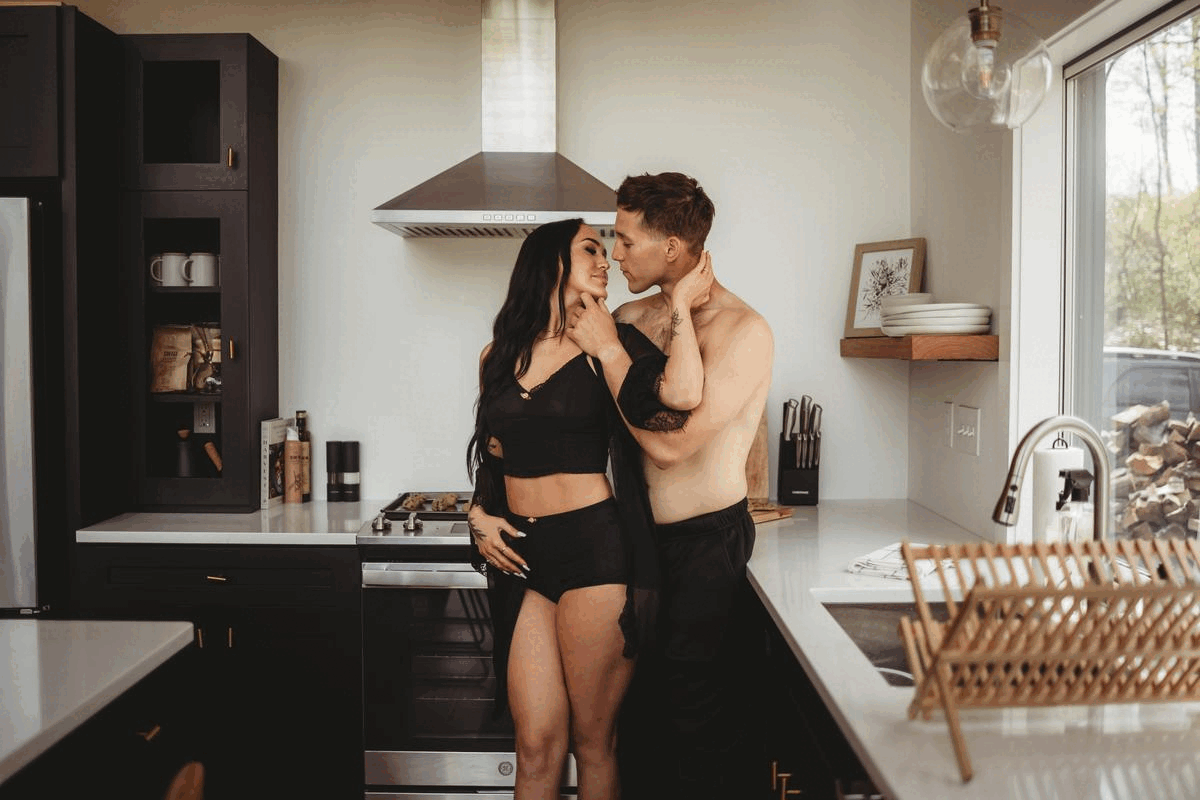 A shirtless man and a woman in black lingerie embracing and looking into each other's eyes in a modern kitchen.