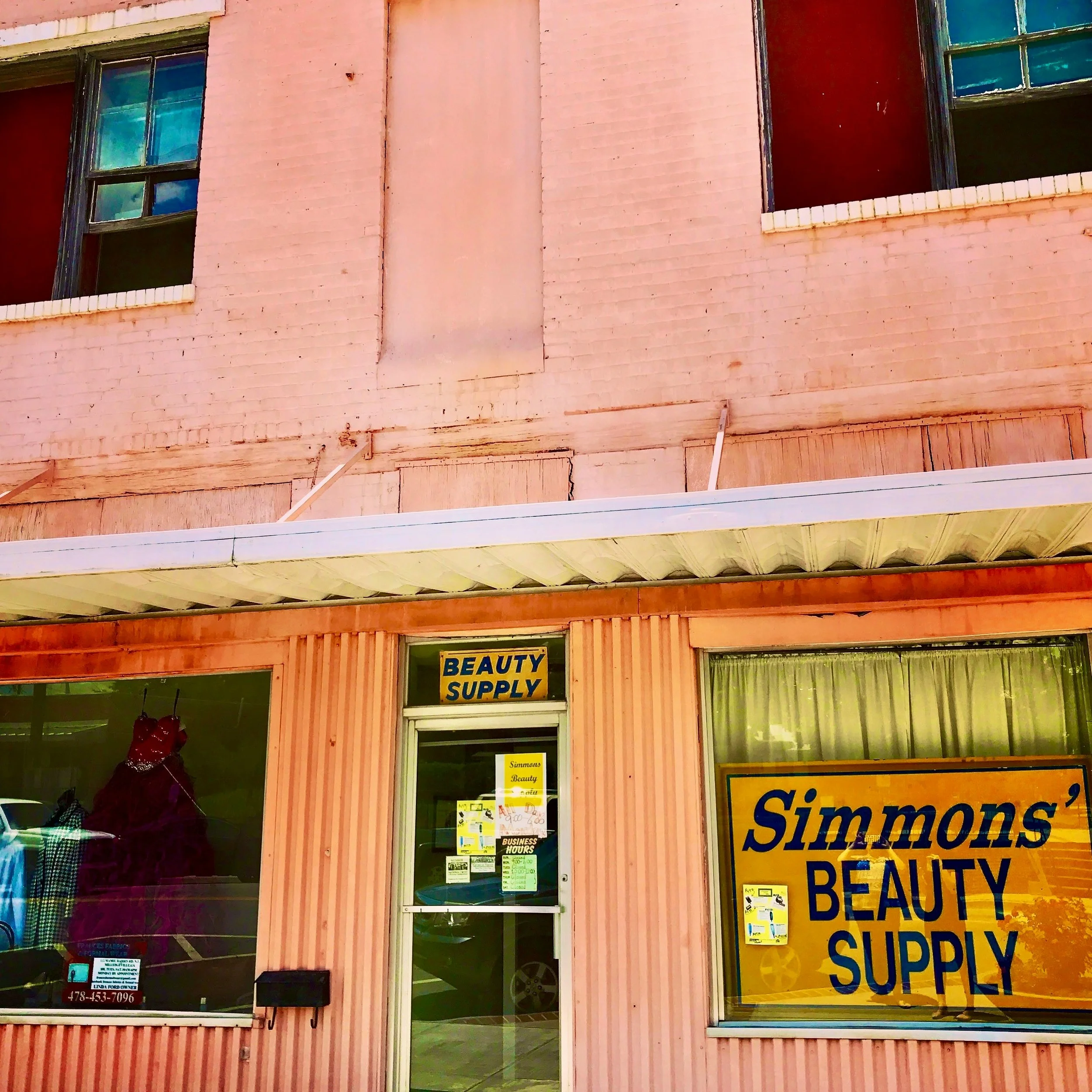 The storefront of a beauty supply store with a pink exterior, a door with a glass window, and large advertising signs indicating beauty supplies and salon hours.