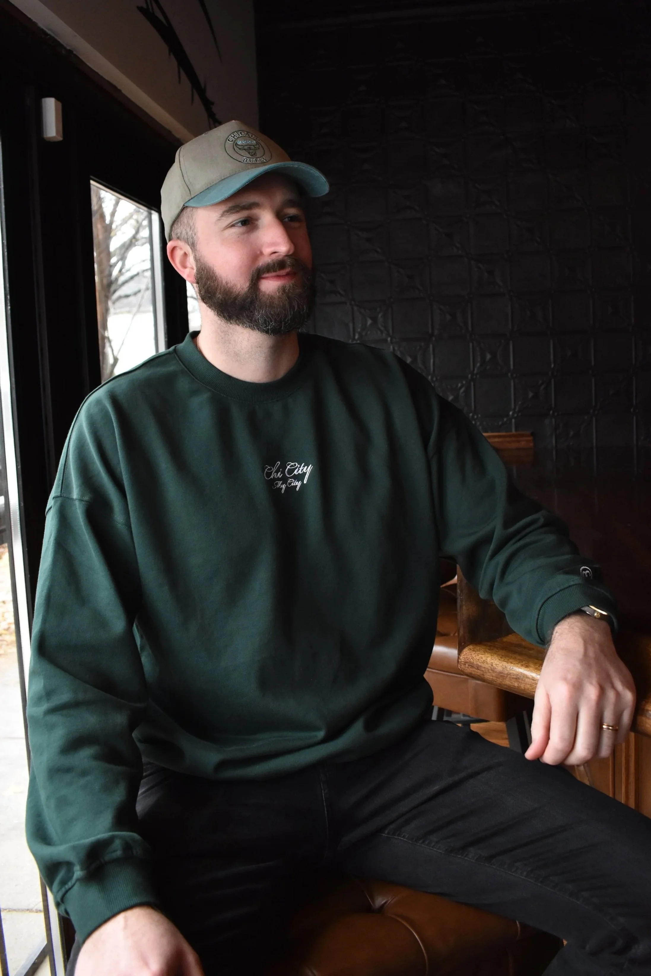 A man wearing a dark green sweatshirt with the text 'Chi City' embroidered on the chest, sitting at a wooden table in a dimly lit indoor setting.