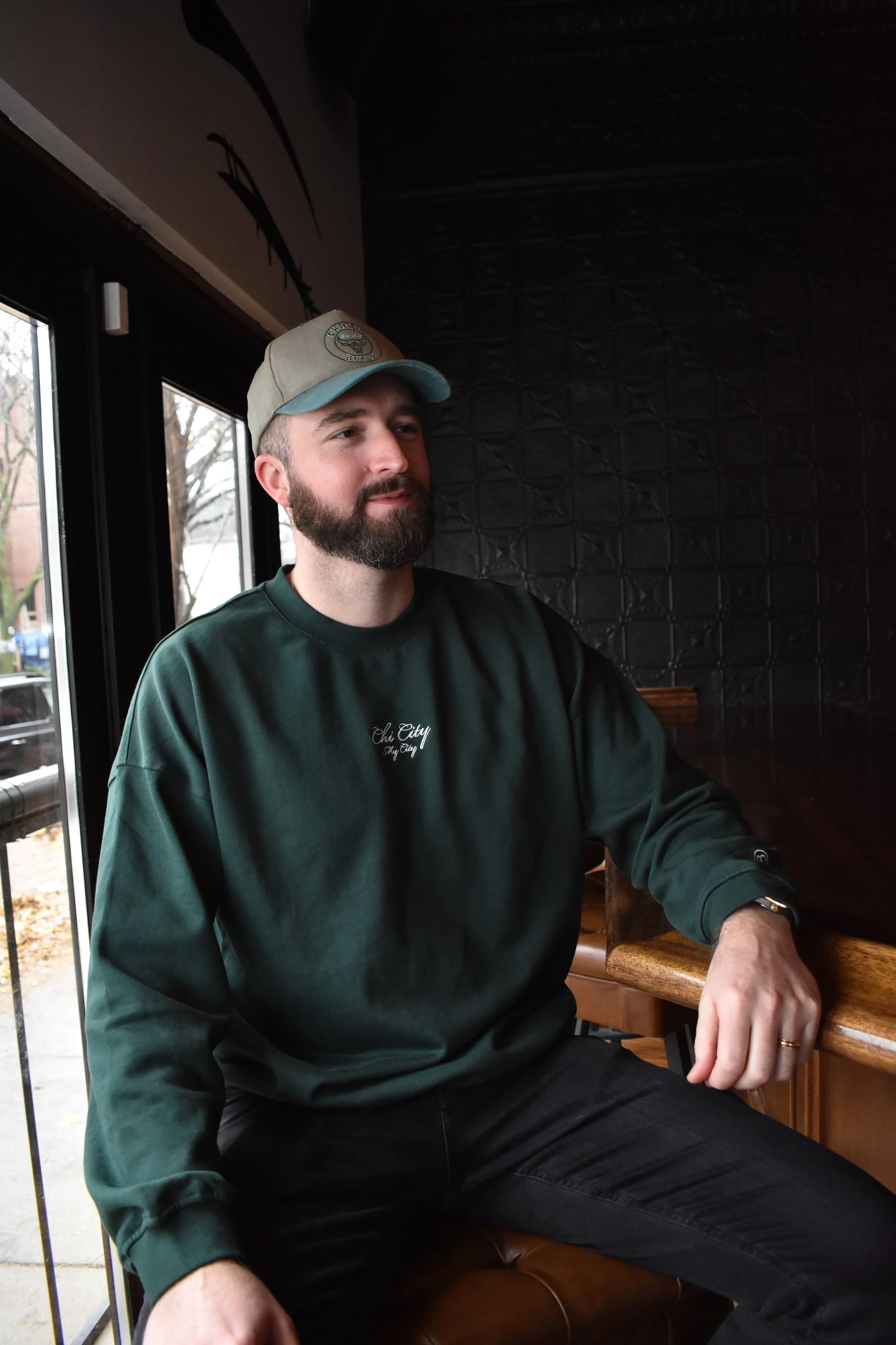 A man wearing a dark green sweatshirt with the text 'Chi City' embroidered on the chest, sitting at a wooden table in a dimly lit indoor setting.