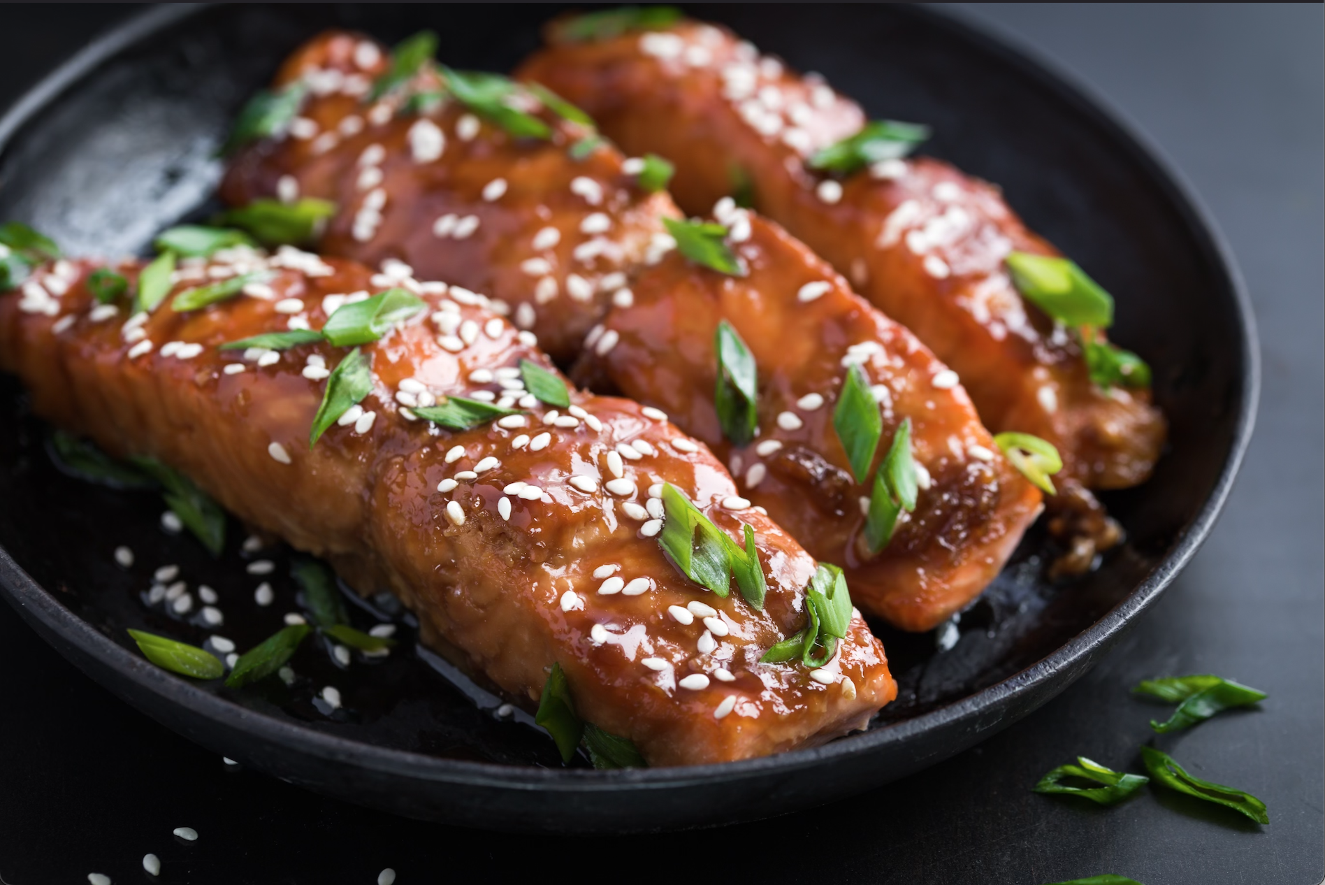 Close-up of three glazed salmon fillets garnished with chopped green onions and sesame seeds in a black skillet at Bettina Cafe Phoenicia New York in the Catskills