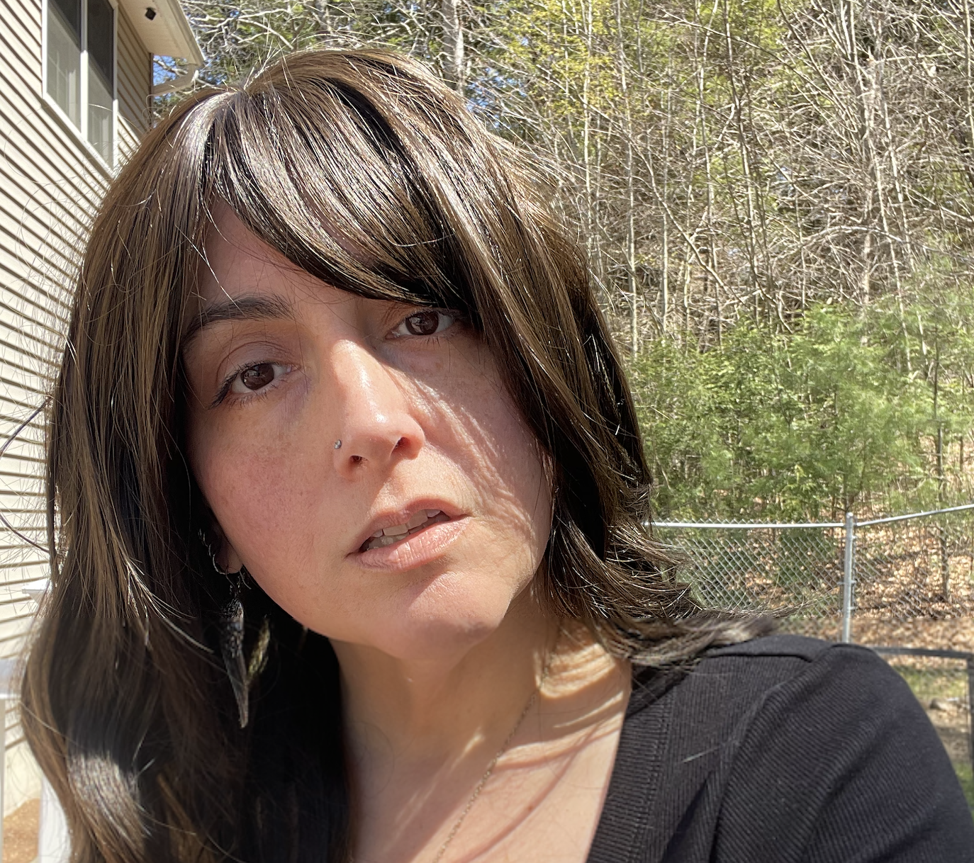 A headshot of a woman with long, brown hair and brown eyes, standing outside with trees behind her.