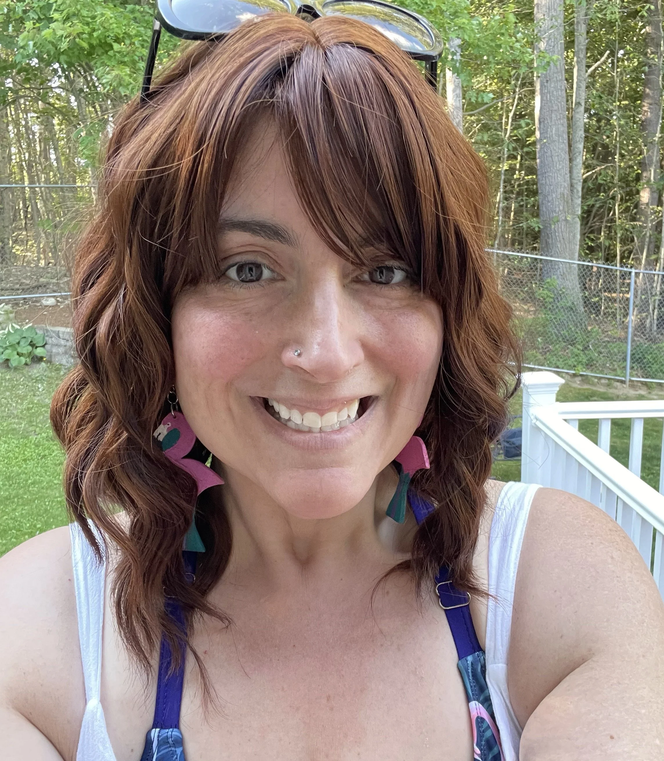Photo of a caucasian woman with shoulder-length, wavy, auburn hair. She is smiling, and had large flamingo earrings.