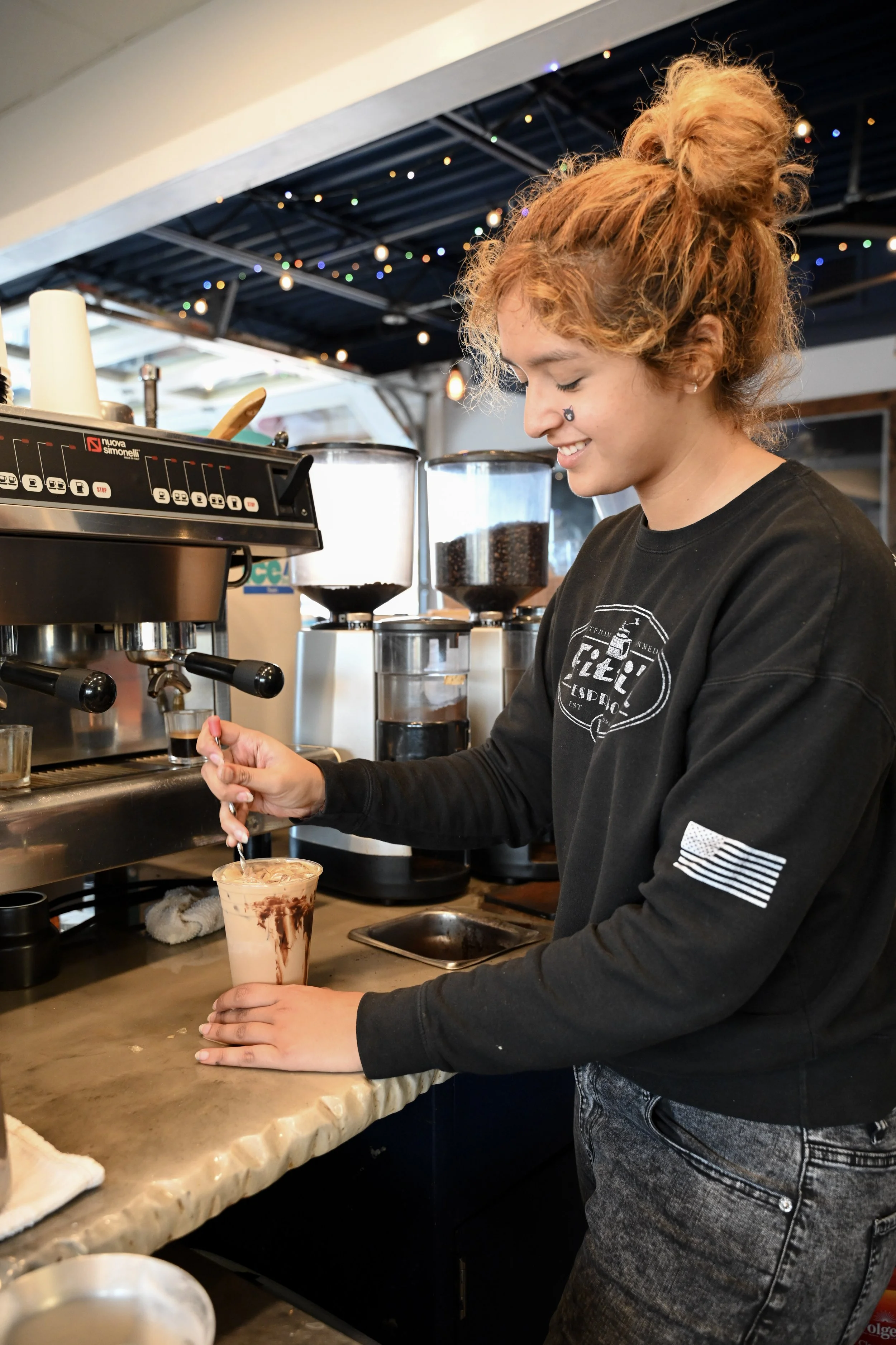 A young woman at Fitti's Espresso smiling as she prepares a coffee drink behind a counter at a coffee shop, with an espresso machine and coffee grinders visible in the background.
