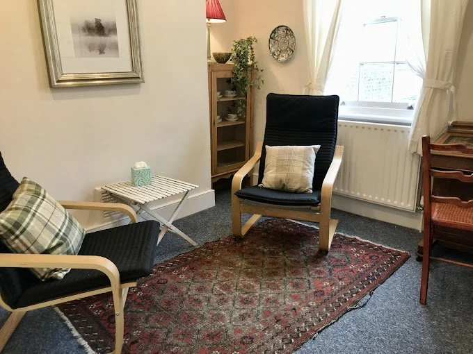 Living room corner with two armchairs, a small side table, a wooden cabinet, a wall clock, a potted plant, and a window with curtains.