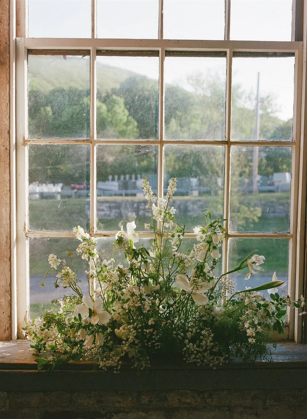 A window with nine small panes and a wooden frame, showing a blurred outdoor scene with trees and a cloudy sky. A bouquet of white flowers is placed on the windowsill inside.