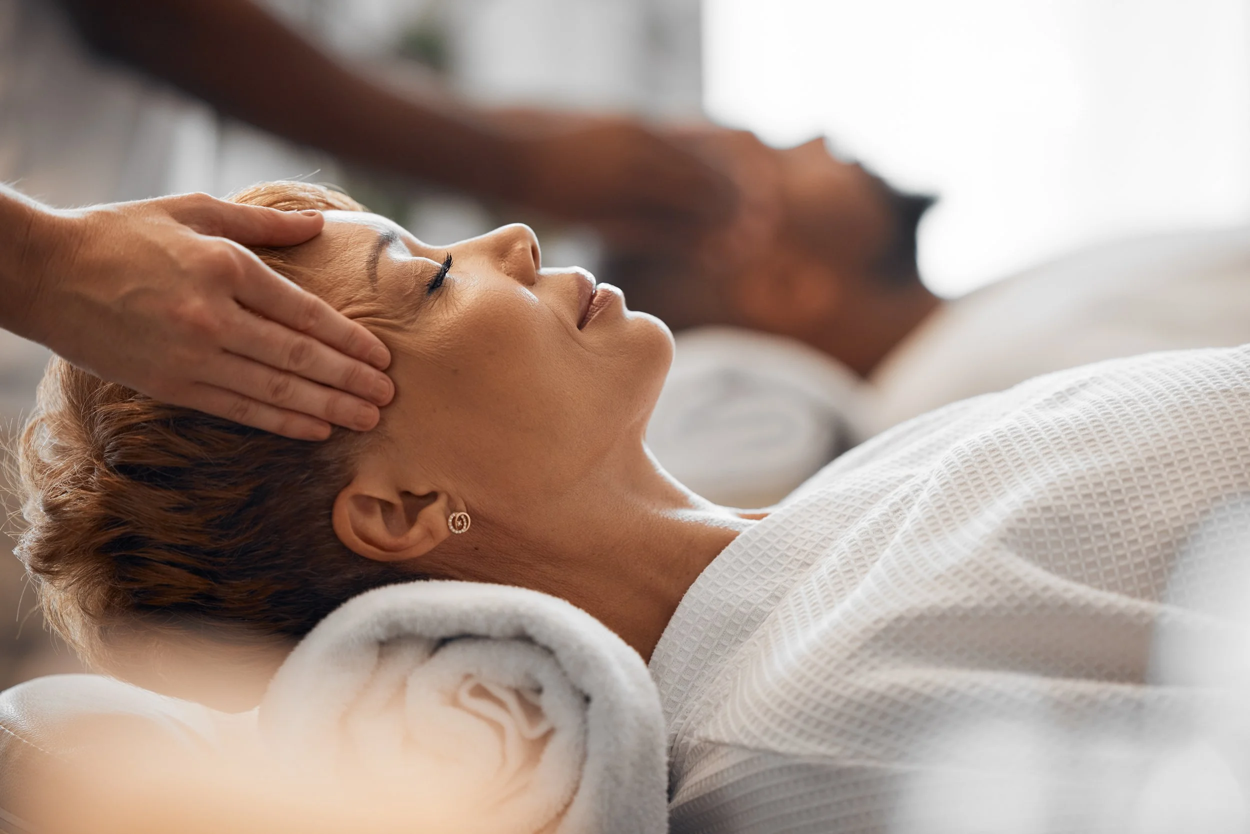 A woman receiving a head massage or facial treatment while lying on a spa bed with her eyes closed, covered with a white towel, and a massage therapist's hands on her forehead.