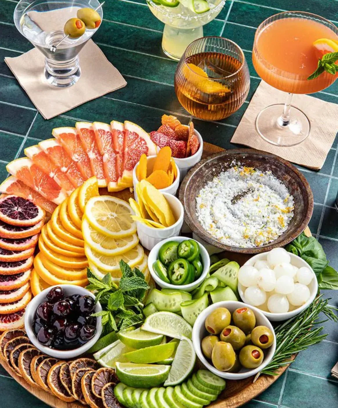 Colorful arrangement of various sliced fruits, olives, lime wedges, and a cheese spread on a wooden platter with drinks in the background.