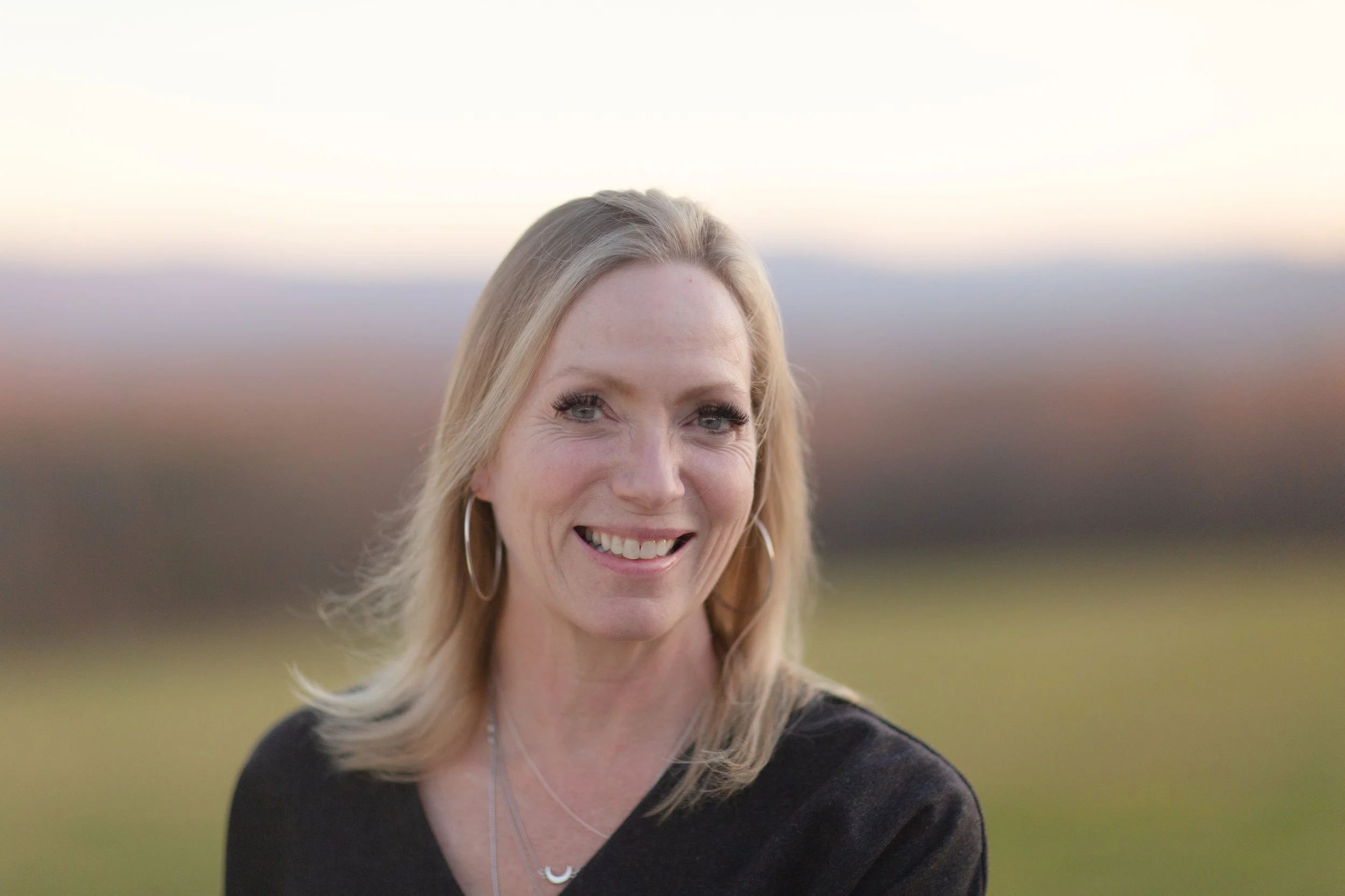 A woman with blonde hair smiling outdoors with a blurred landscape background at sunset.