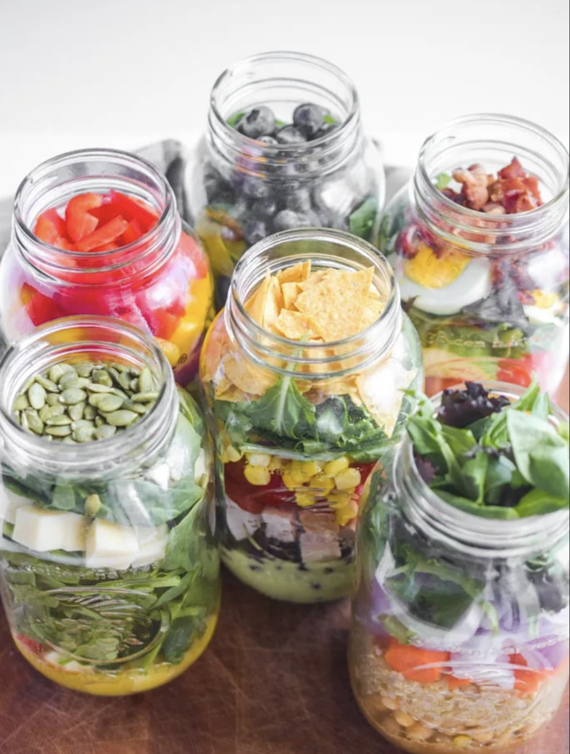 Six glass jars filled with various layered salads containing vegetables, cheese, and toppings on a wooden surface.
