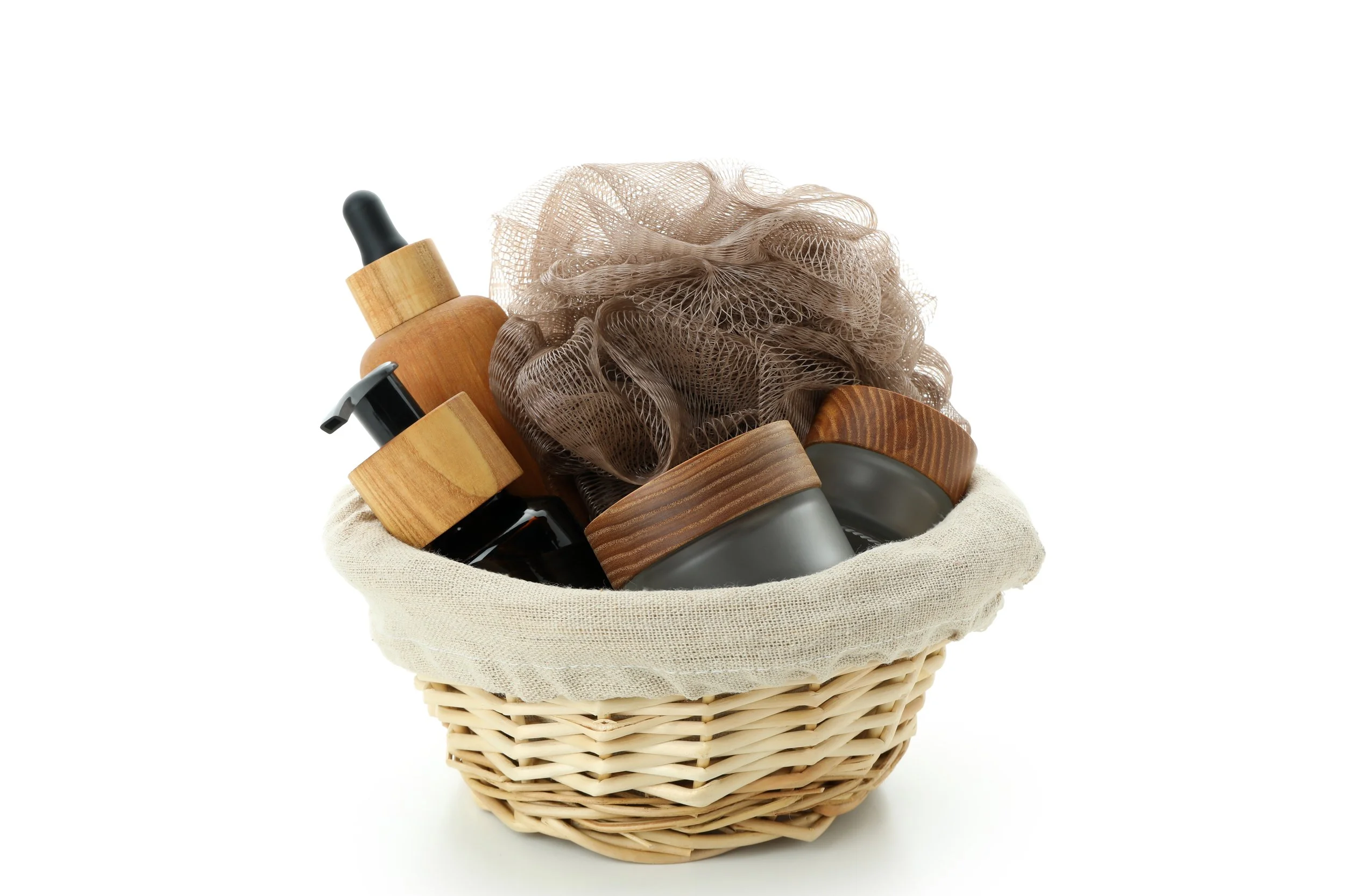 A gift basket containing brown and black bottles with pump tops, a mesh bath sponge, and black containers with wooden lids, all arranged inside a woven basket lined with a fabric cloth.