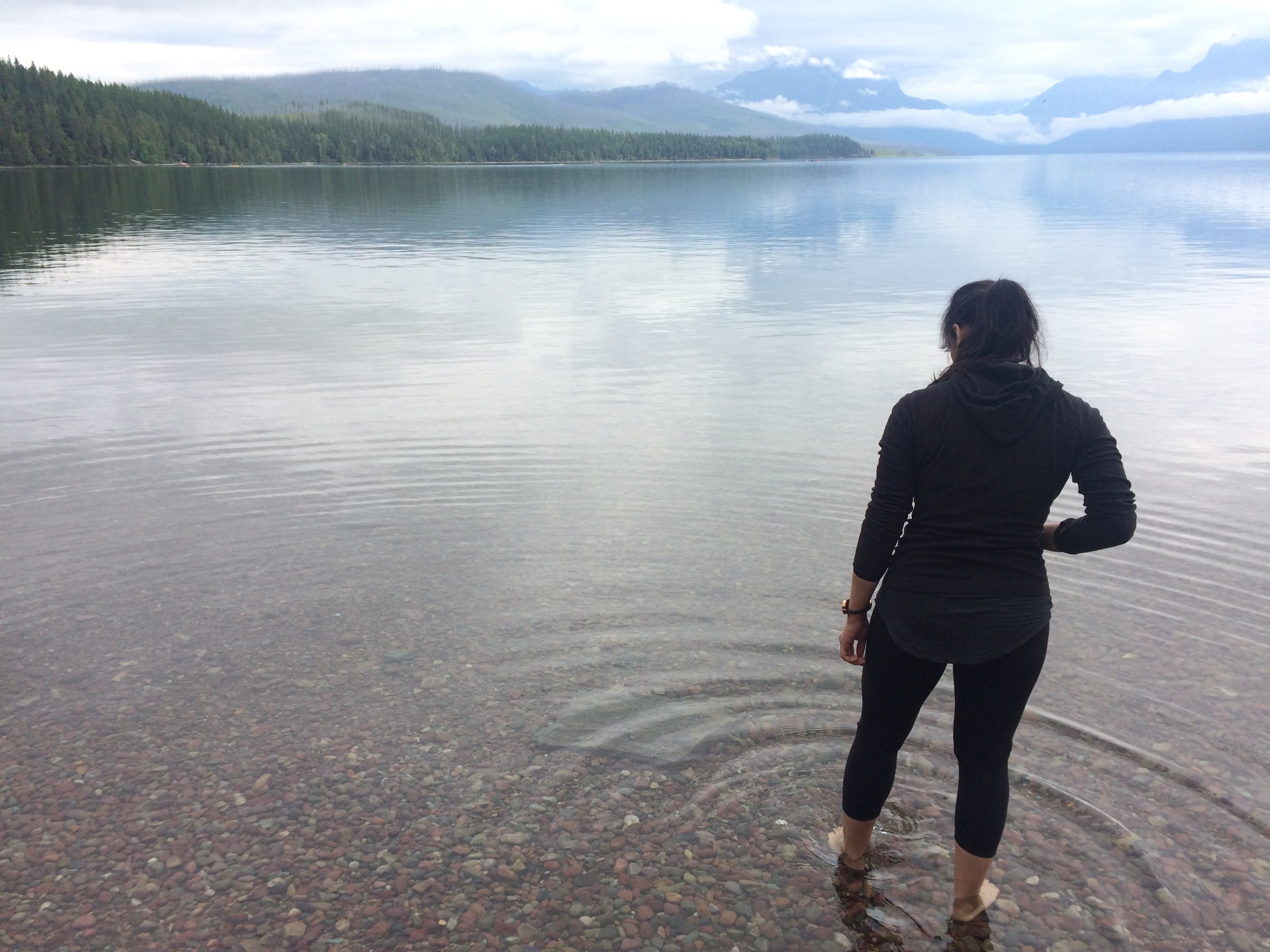 Person wearing a black hoodie and black pants standing in shallow water at the edge of a calm lake with mountains and fog in the background.