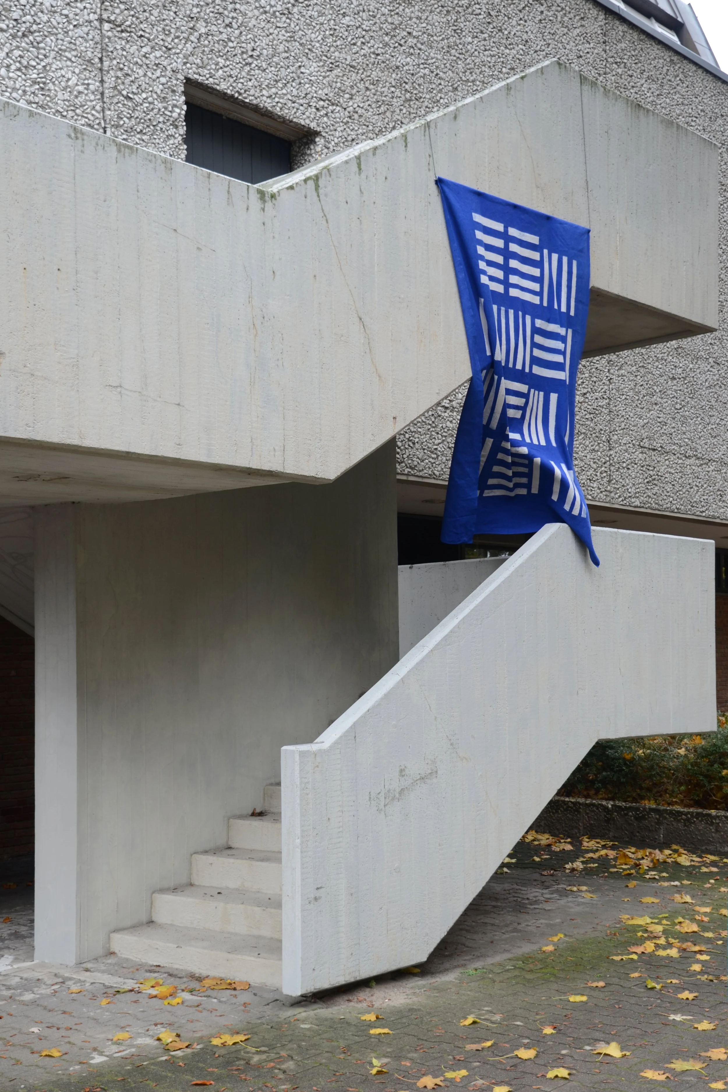 Ein Betontreppe in eines Gebäudes mit einer blauen Fahne, die an der Wand hängt, und Herbstblätter auf dem Boden, Blaudruck von Franziska Lutze.