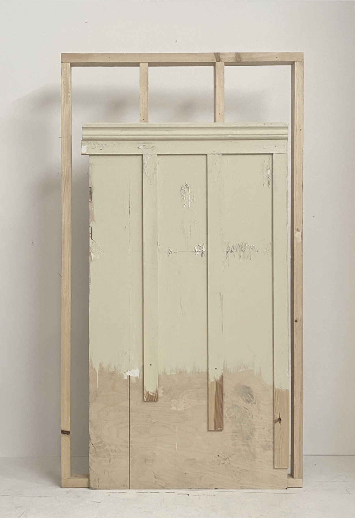 Ein Holzrahmen, in dem eine alte, cremefarbene Holztür mit abblätternder Farbschicht angebracht ist, steht vor einer weißen Wand.