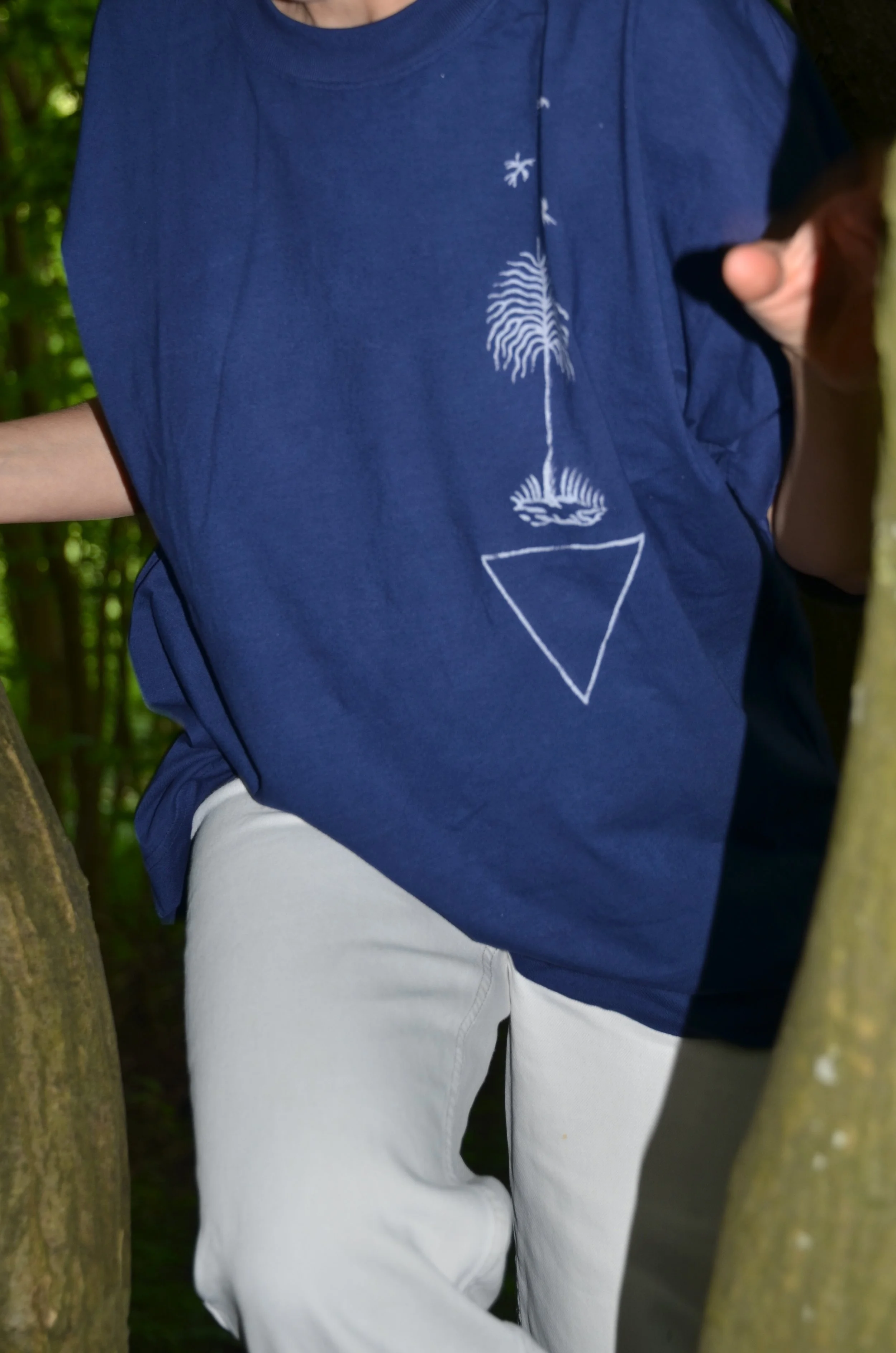 Nahaufnahme einer Person, die in einem Wald zwischen Bäumen sitzt, trägt ein dunkelblaues T-Shirt mit einer weißen Baum- und Dreieckgestaltung, kombiniert mit weißen Hosen. T-Shirt von Franziska Lutze in Kollaboration mit A.D.Deertz.