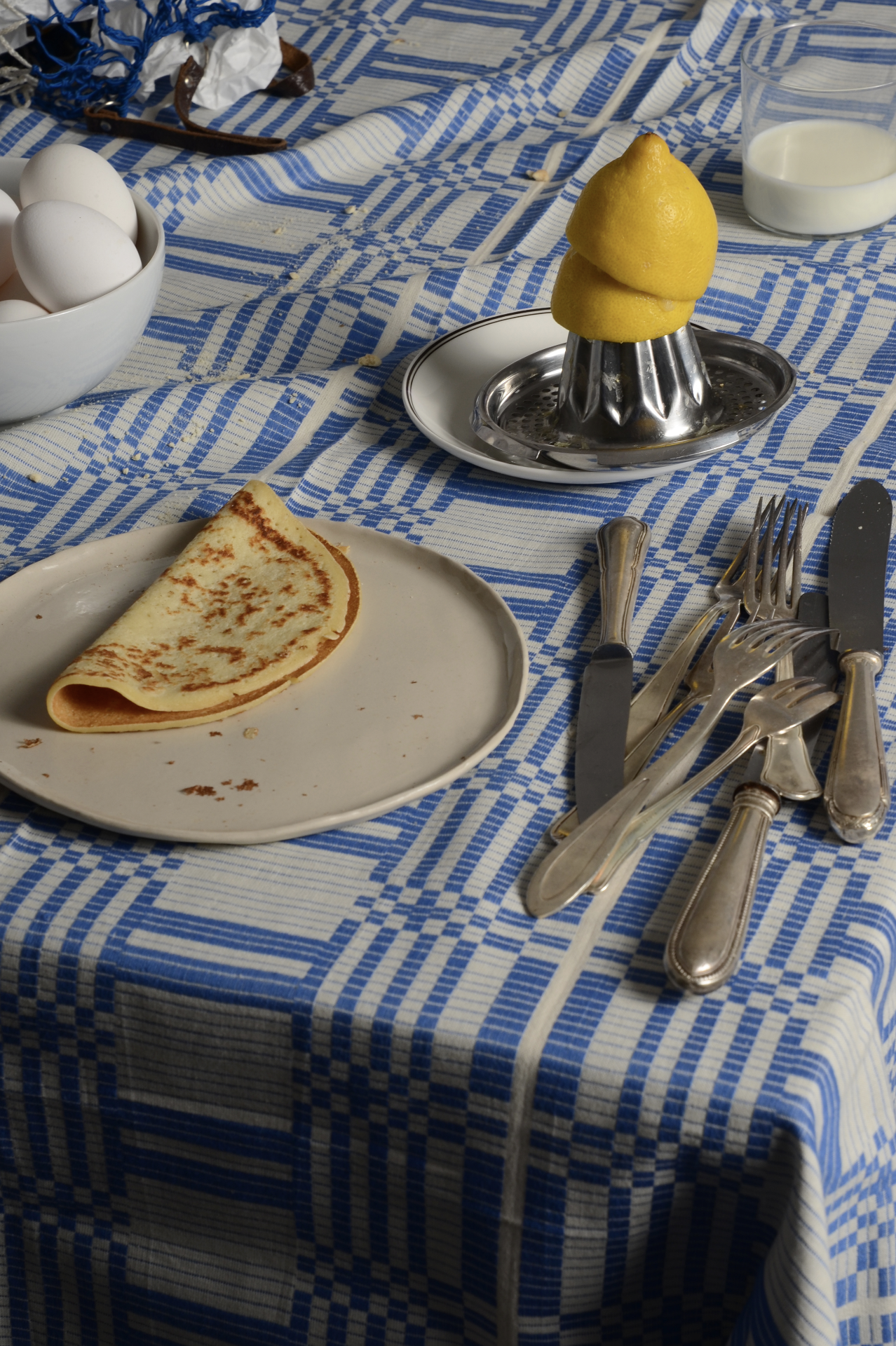 Ein blauer und weißer Tischläufer mit Mahlzeiten, darunter ein Teller mit einem Pfannkuchen, ein Glas Milch, Eier in einer Schüssel, eine Zitrone auf einer Metallpresse, und Besteck