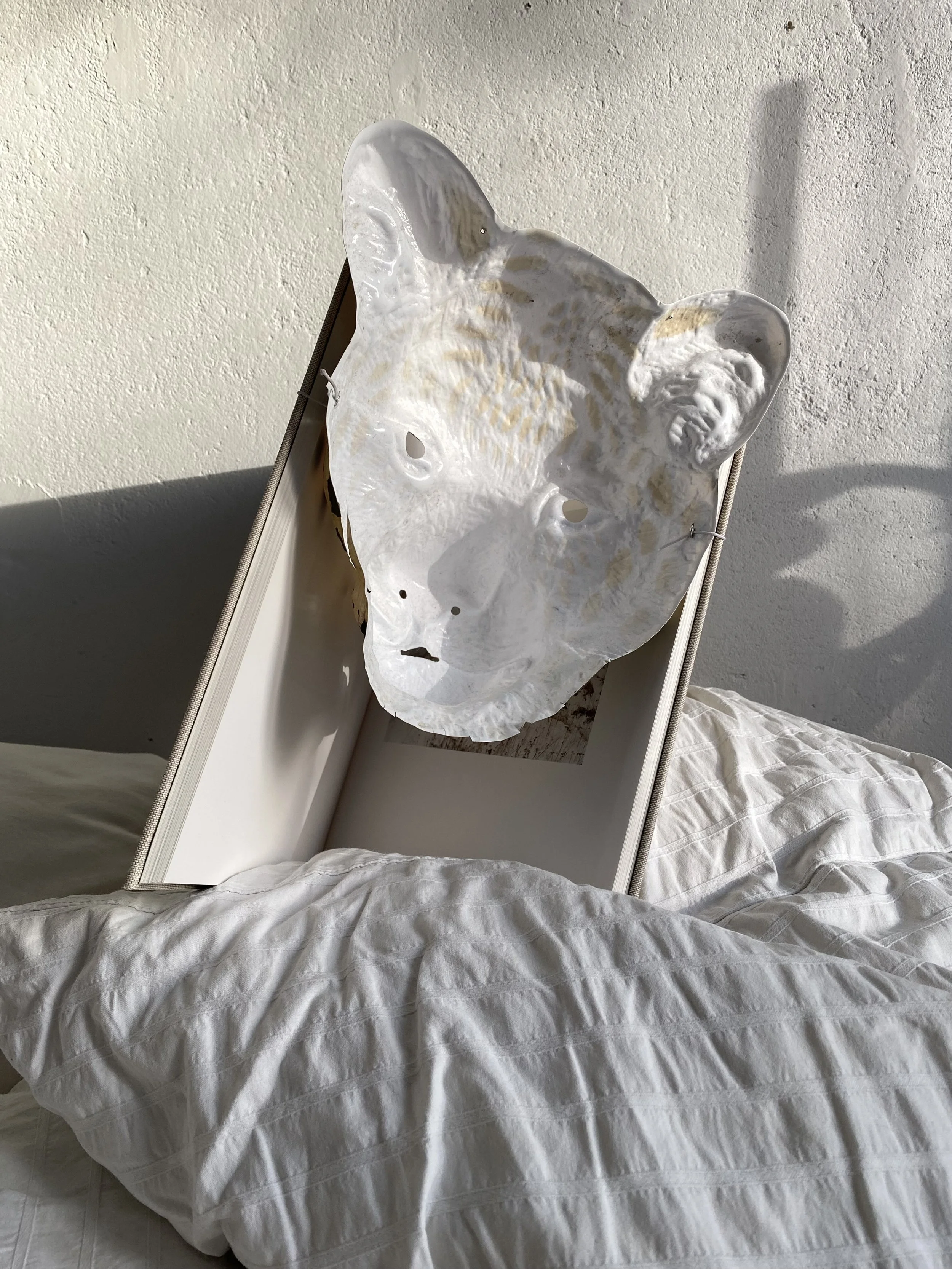 Ein Bett mit weißen Bettwäsche und einem weißen Nachttisch. Auf dem Nachttisch steht eine große weiße Maske eines Tierkopfes, wahrscheinlich eines Löwen.