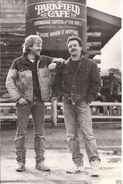 John Varian and Steve Johnson standing in front of the Parkfield Cafe sign, smiling and talking. They are wearing casual jackets and jeans.