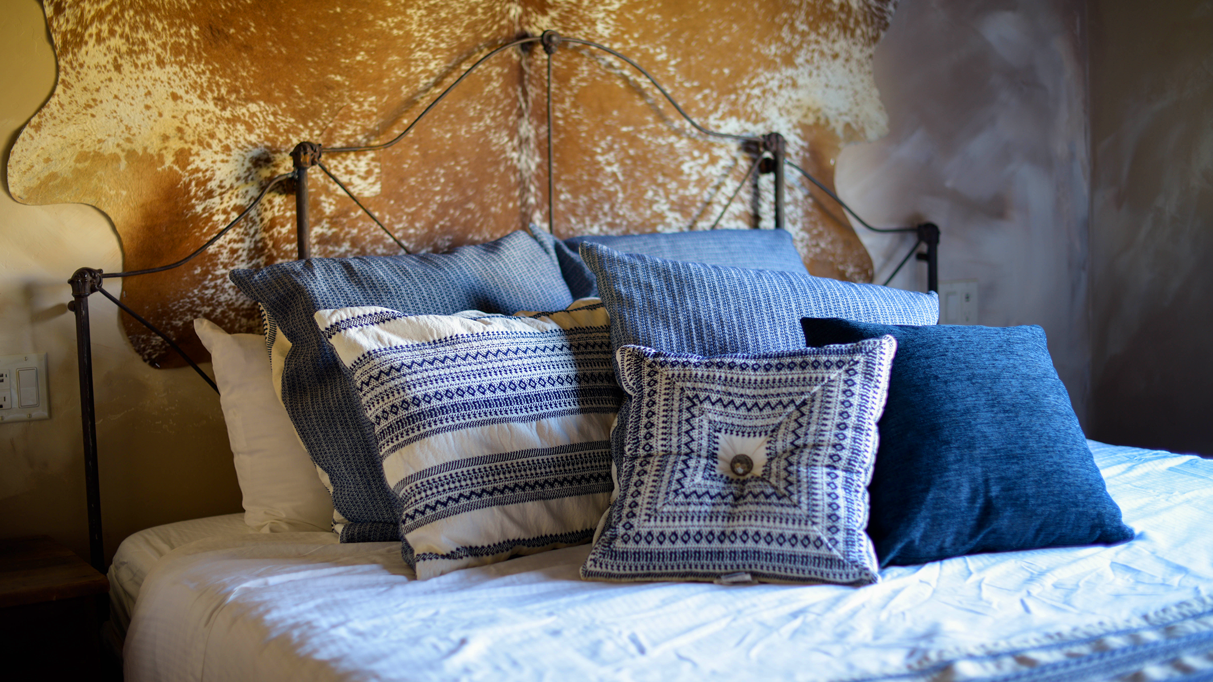 A cozy bed with several blue and white patterned pillows, antique iron headboard, set against a cowhide hung on the wall in the historic Post Office Room at the Parkfield Lodge on the Central Coast of California.
