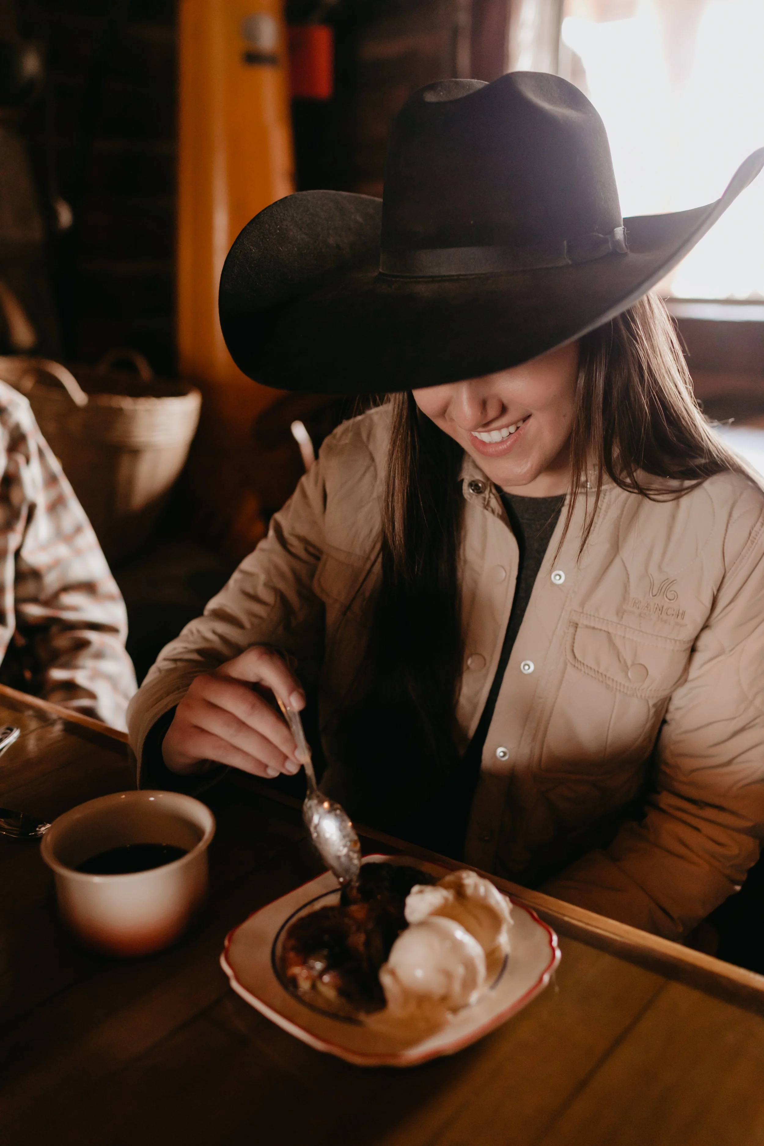 A cowgirl in a cowboy hat and a beige jacket is smiling while eating a Parkfield Cafe Apple Dumpling dessert with ice cream, sitting at a wooden table in the cozy, rustic Parkfield Cafe on California's Central Coast.
