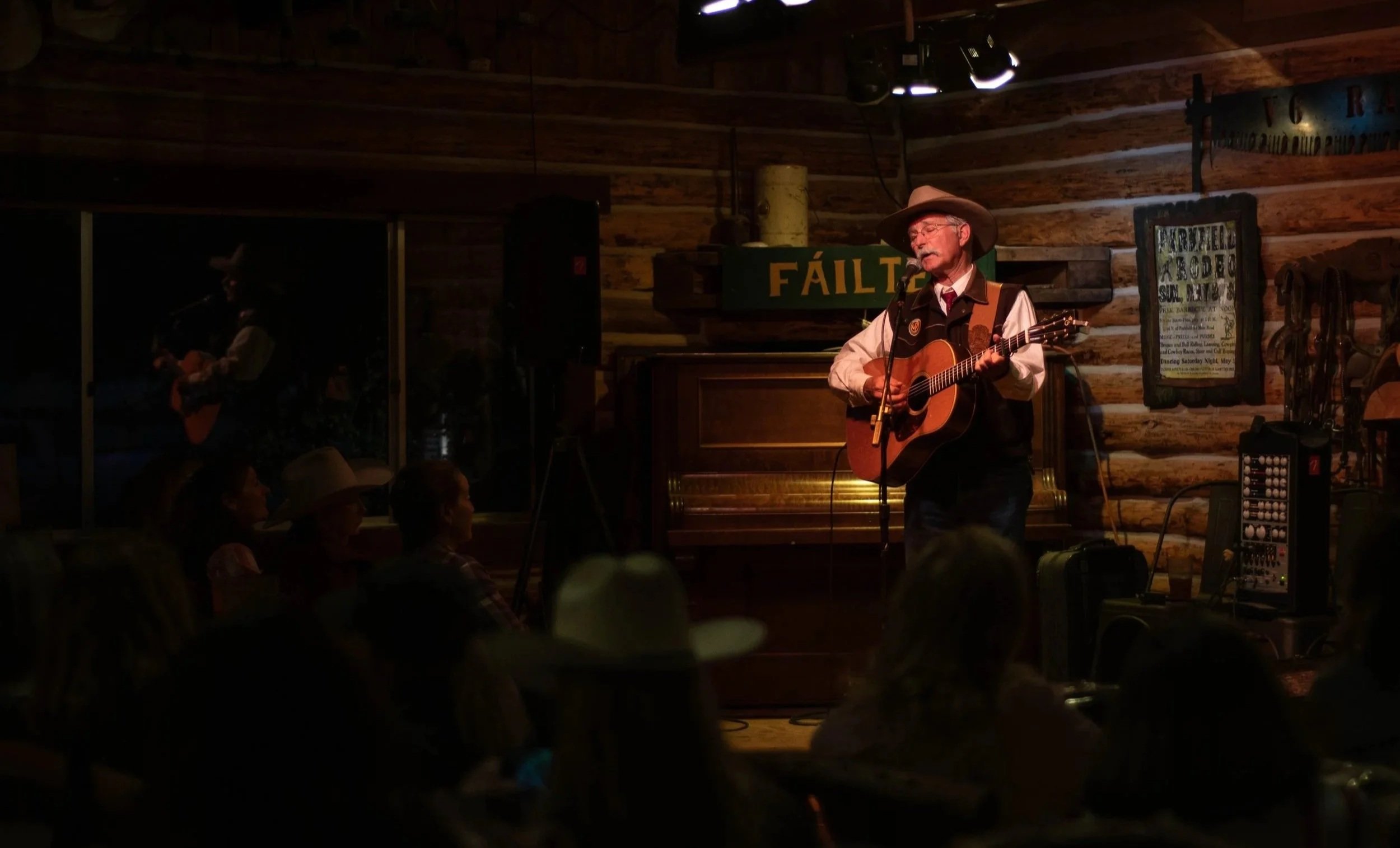 Dave Stamey playing an acoustic guitar and singing into a microphone on stage in the rustic log cabin the Parkfield Cafe, with an audience watching.