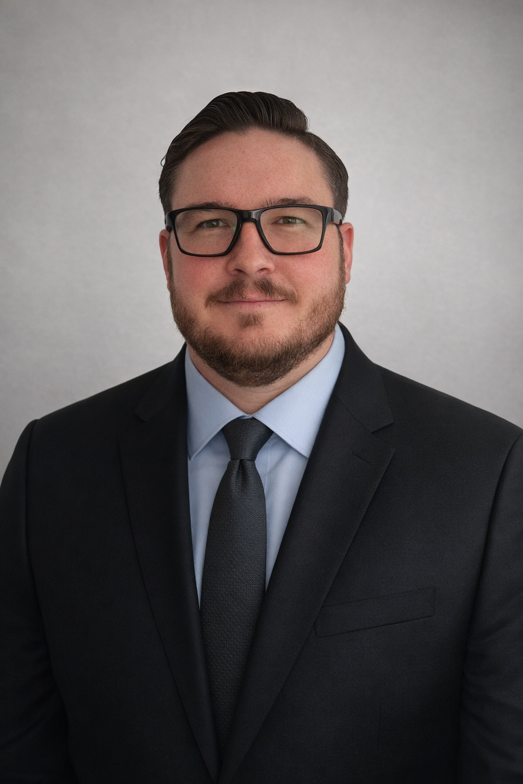 A man with glasses, facial hair, and a slight smile, dressed in a black suit, light blue shirt, and dark tie, standing against a plain gray background.