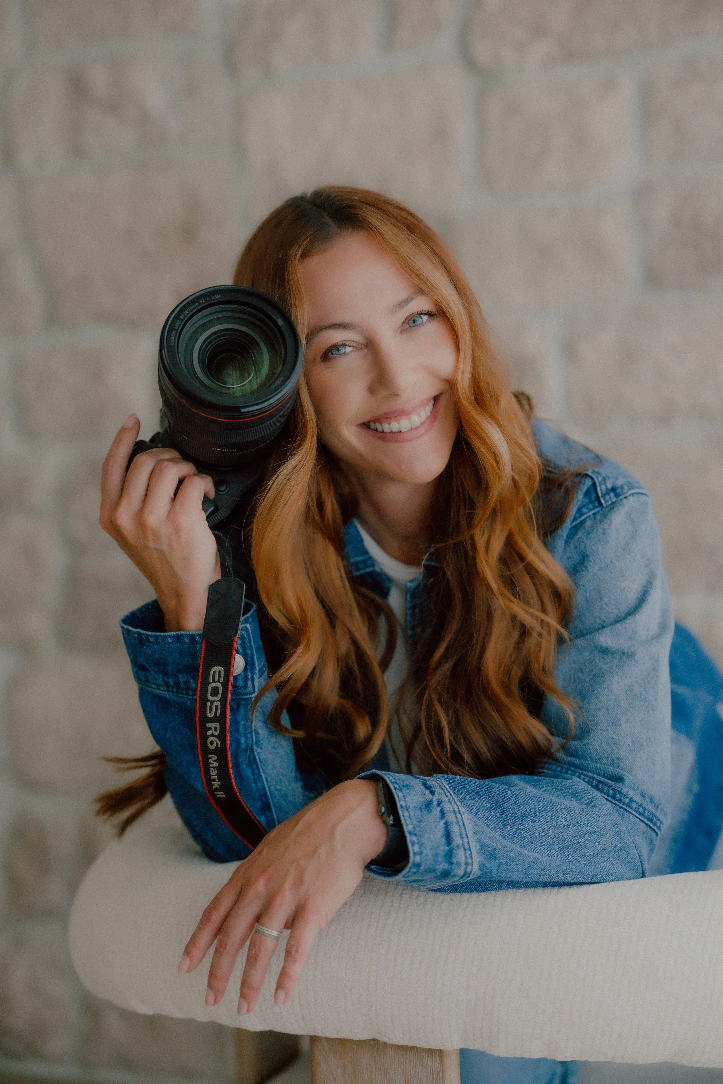 A woman with long red hair, blue eyes, and a smile, holding a camera lens to her eye. She is wearing a denim jacket and is seated, with a light-colored textured surface and a stone wall background.