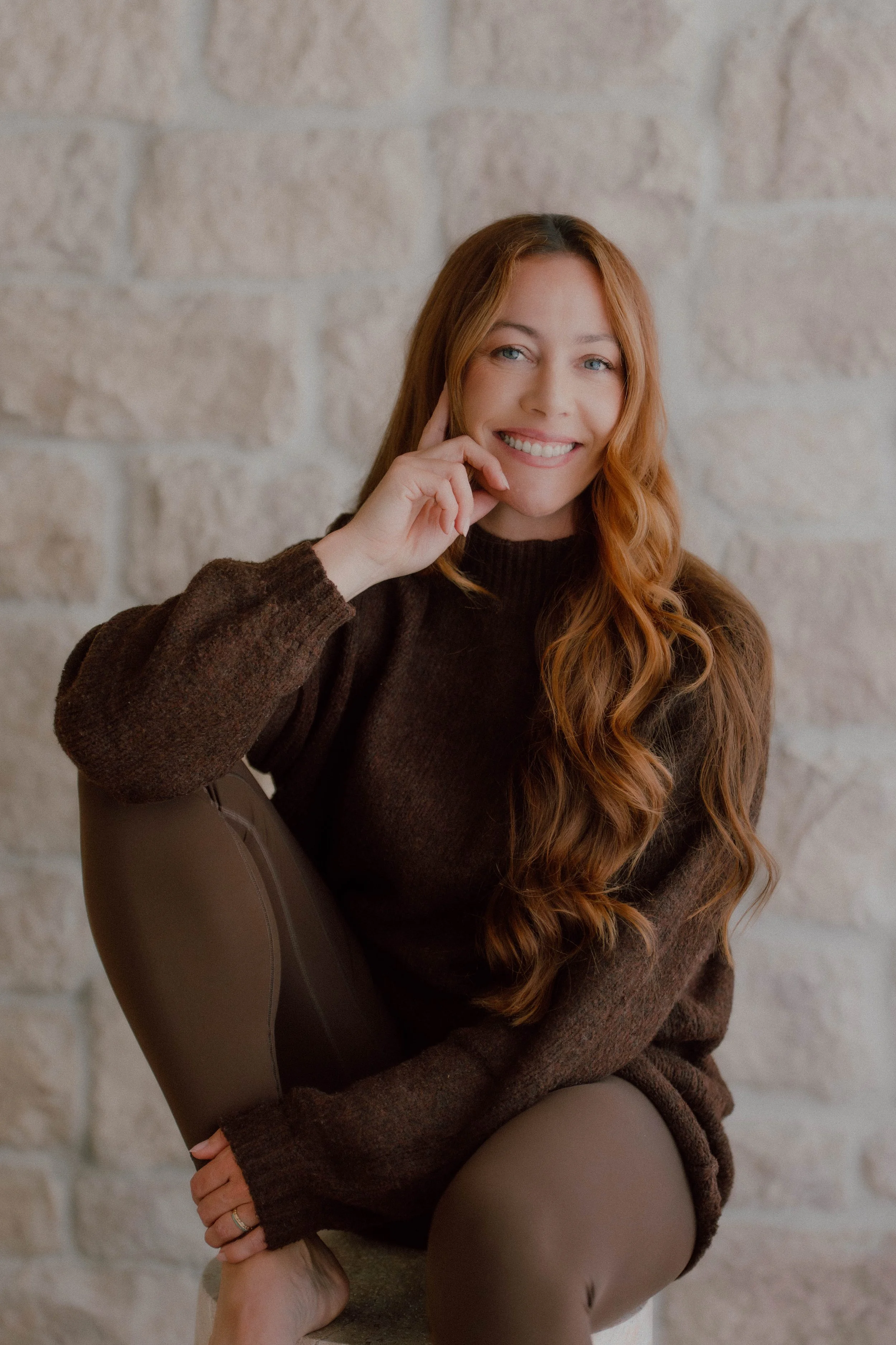 A smiling woman with long red hair sitting in front of a light-colored brick wall. She is wearing a dark brown sweater and beige pants, with one hand touching her face and the other resting on her knee.