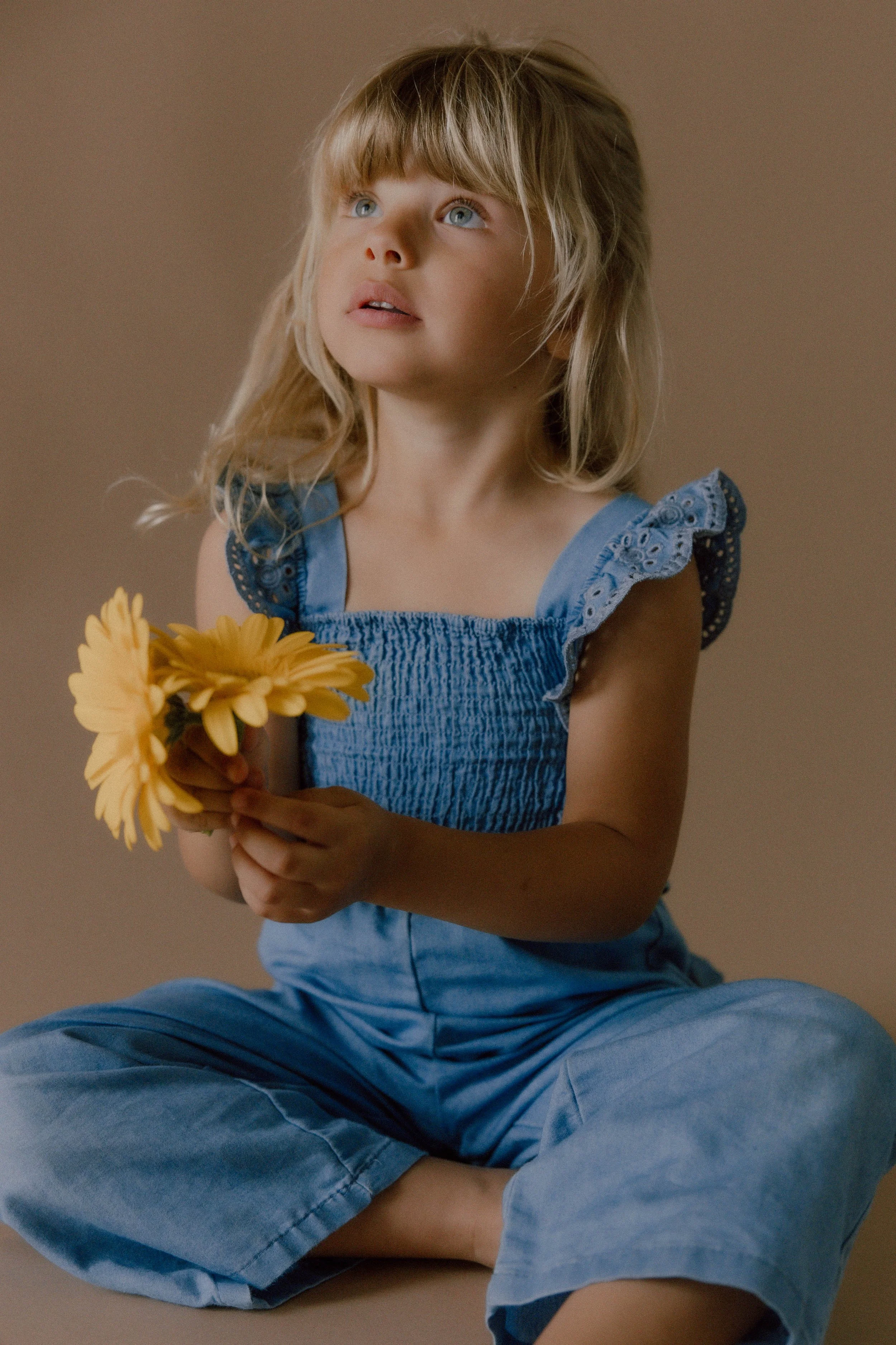 A young girl with blonde hair and blue eyes, sitting cross-legged on the floor, holding a yellow flower and looking thoughtfully upwards.