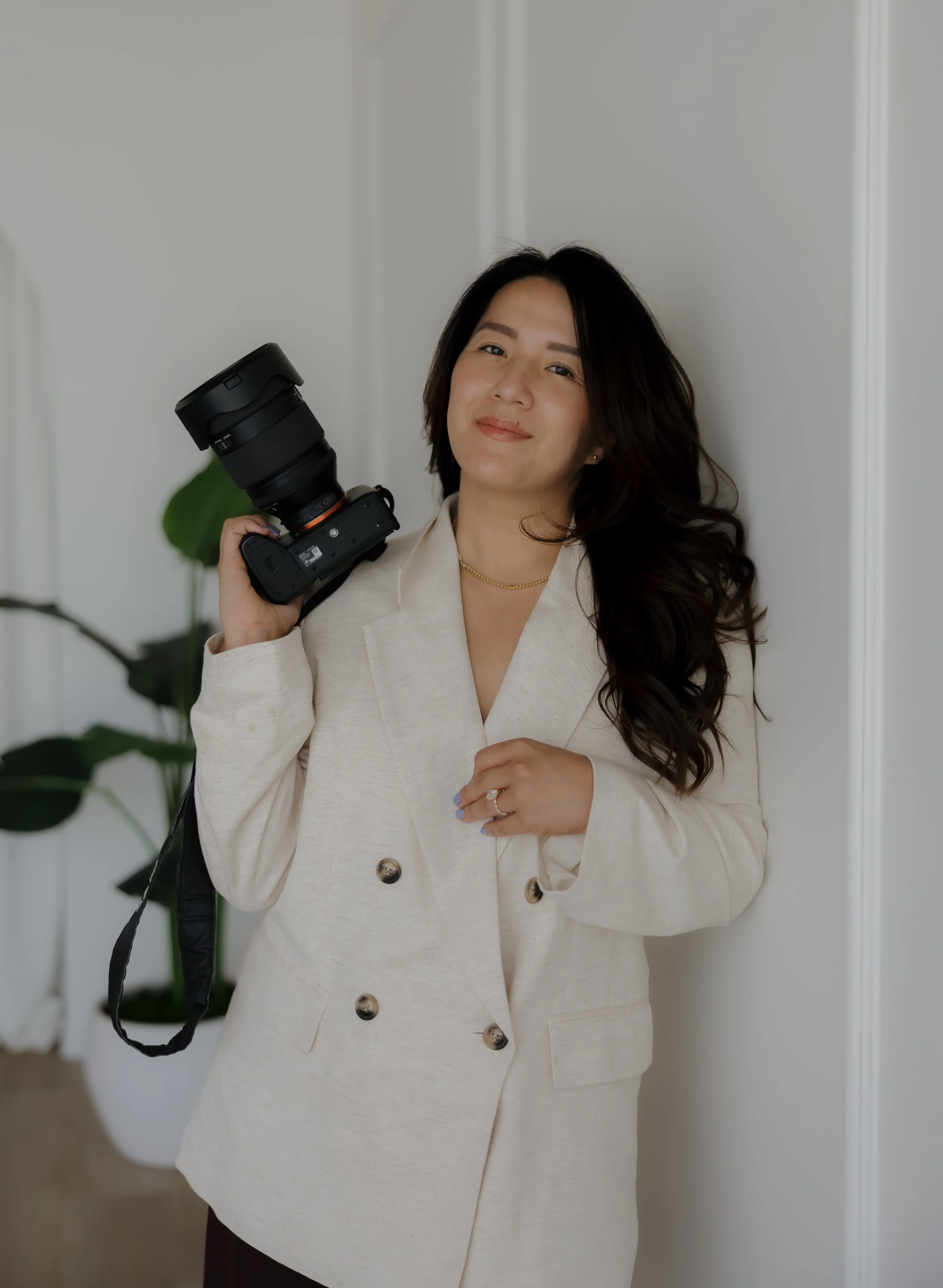A woman with long, wavy black hair wearing a cream-colored blazer and skirt holding a camera in her right hand.