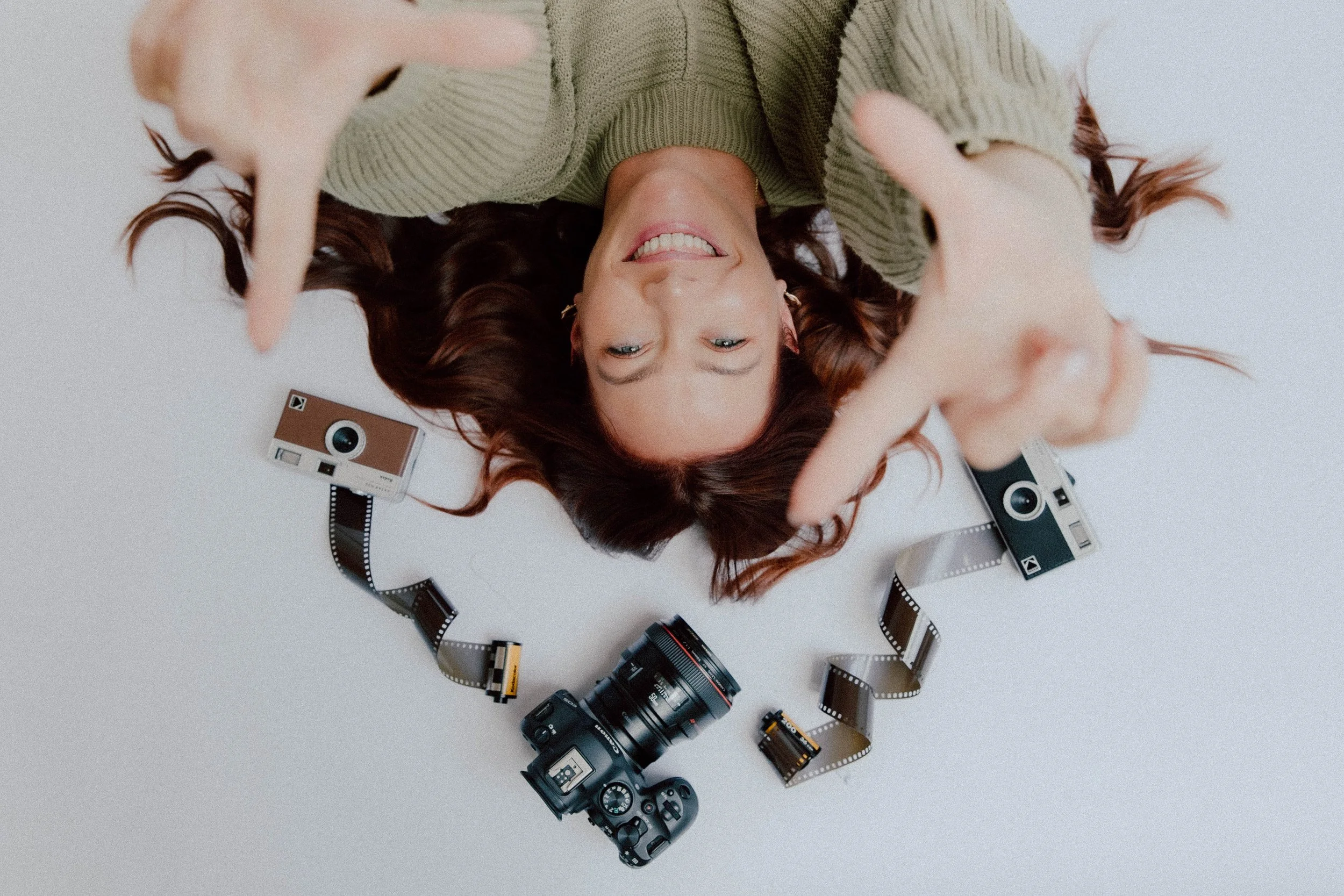 A woman lying on her back smiling, surrounded by vintage cameras, film rolls, and photographic equipment on a white surface.