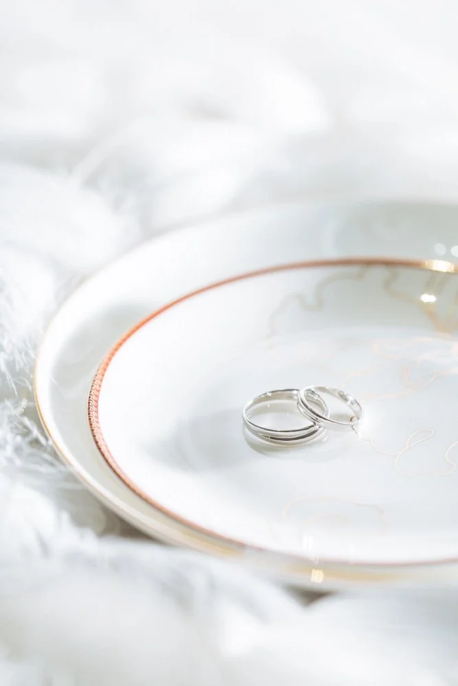 Two wedding rings on a decorative white plate with gold trim, resting on soft white fabric.