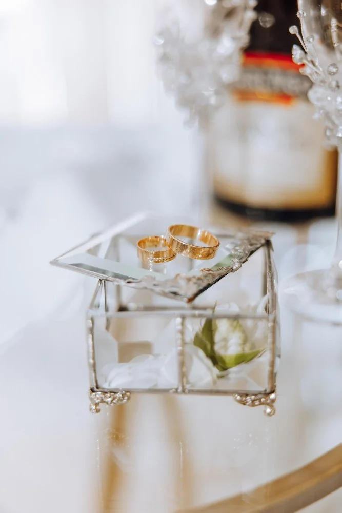 Gold wedding rings on a mirror in a glass jewelry box with a flower inside, placed on a white table.