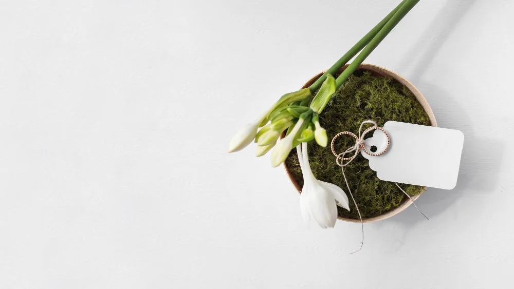 White flowers, possibly lilies, in a small round pot with moss, on a white surface, with a blank white tag and a pair of rings.