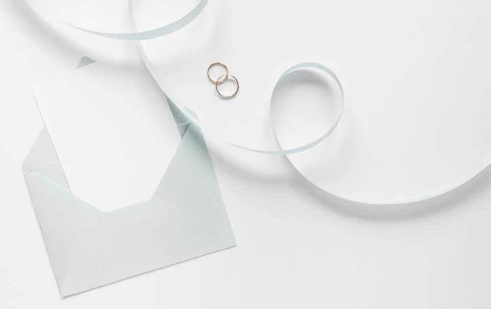 Two gold wedding rings placed on a white surface, next to a blank white envelope and white decorative ribbon.