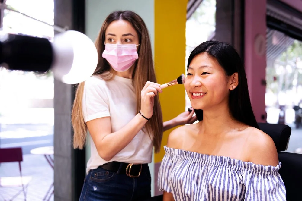Makeup artist applying makeup to a woman sitting in a salon chair, with both smiling. The woman is wearing an off-the-shoulder striped top, and the makeup artist is wearing a pink face mask.