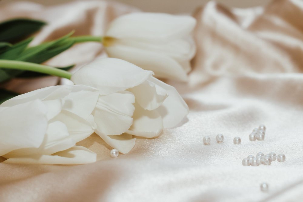 White tulips with green stems and leaves lying on a silky beige fabric, with small white pearls scattered on the fabric.