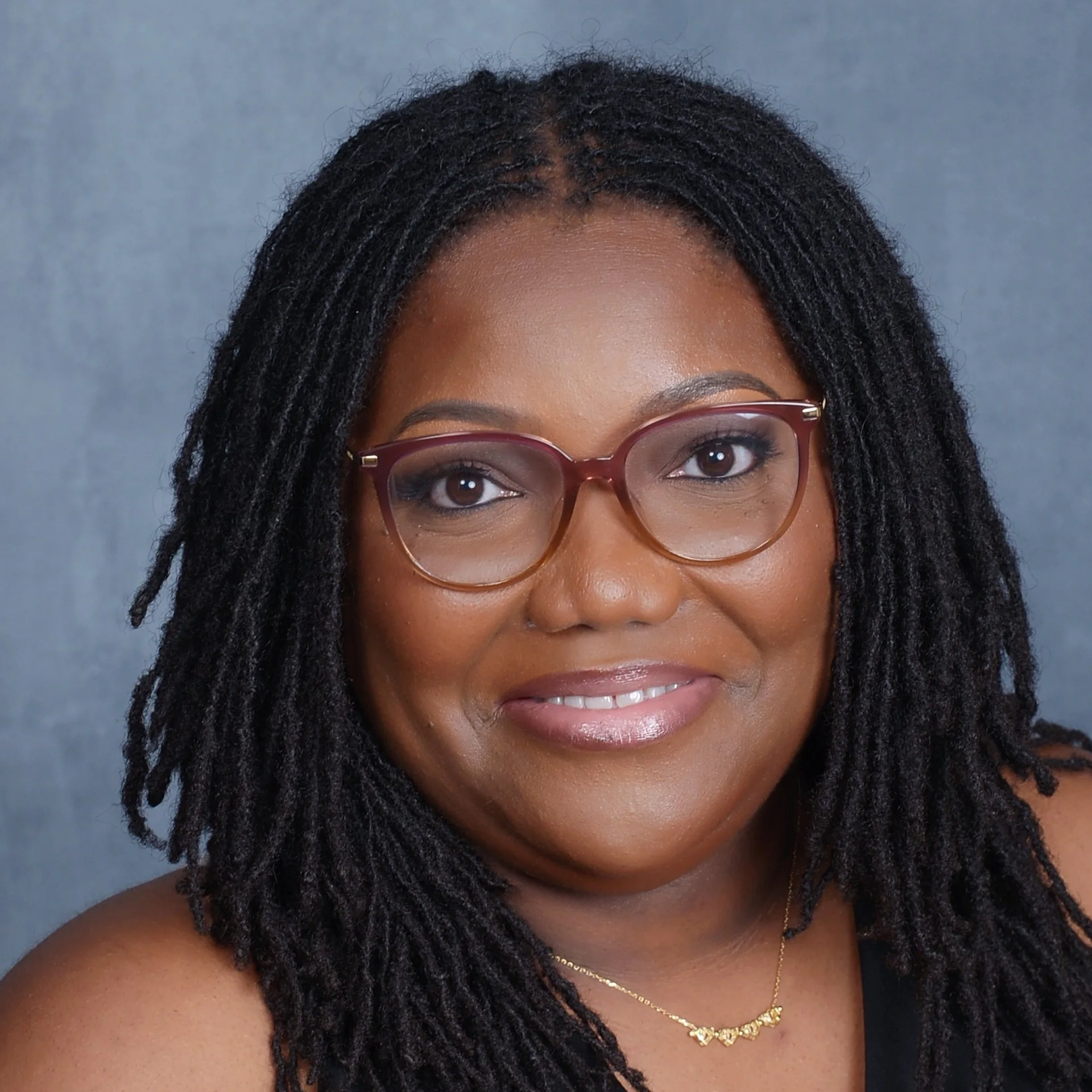 A smiling woman with medium-dark skin, black dreadlocks, wearing glasses, a black top, a gold necklace, and makeup, standing against a grey background.