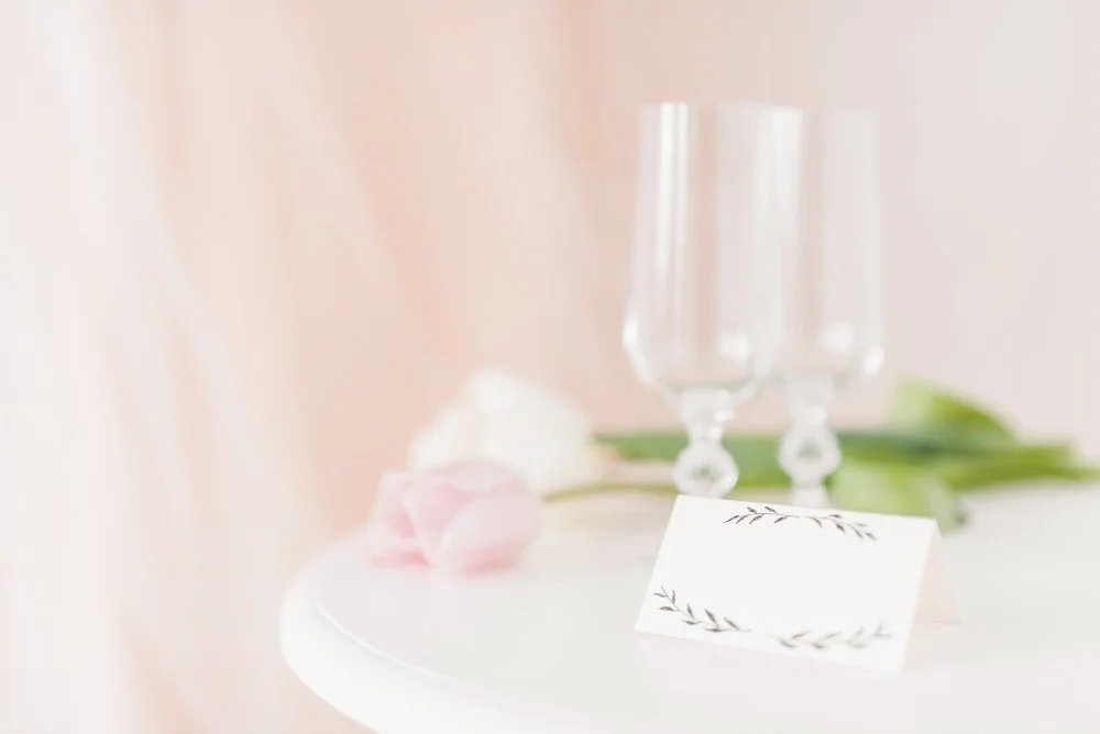 A table setting with empty champagne glasses, pink and white roses, a blank place card, and green leaves, with a soft pink background.