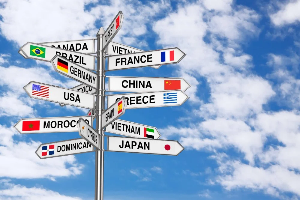 Signpost with flags and country names pointing in various directions, including Canada, Brazil, Germany, USA, Mexico, Cyprus, Dominican Republic, Turkey, Vietnam, France, China, Greece, Spain, Morocco, Japan, and others, against a cloudy sky.