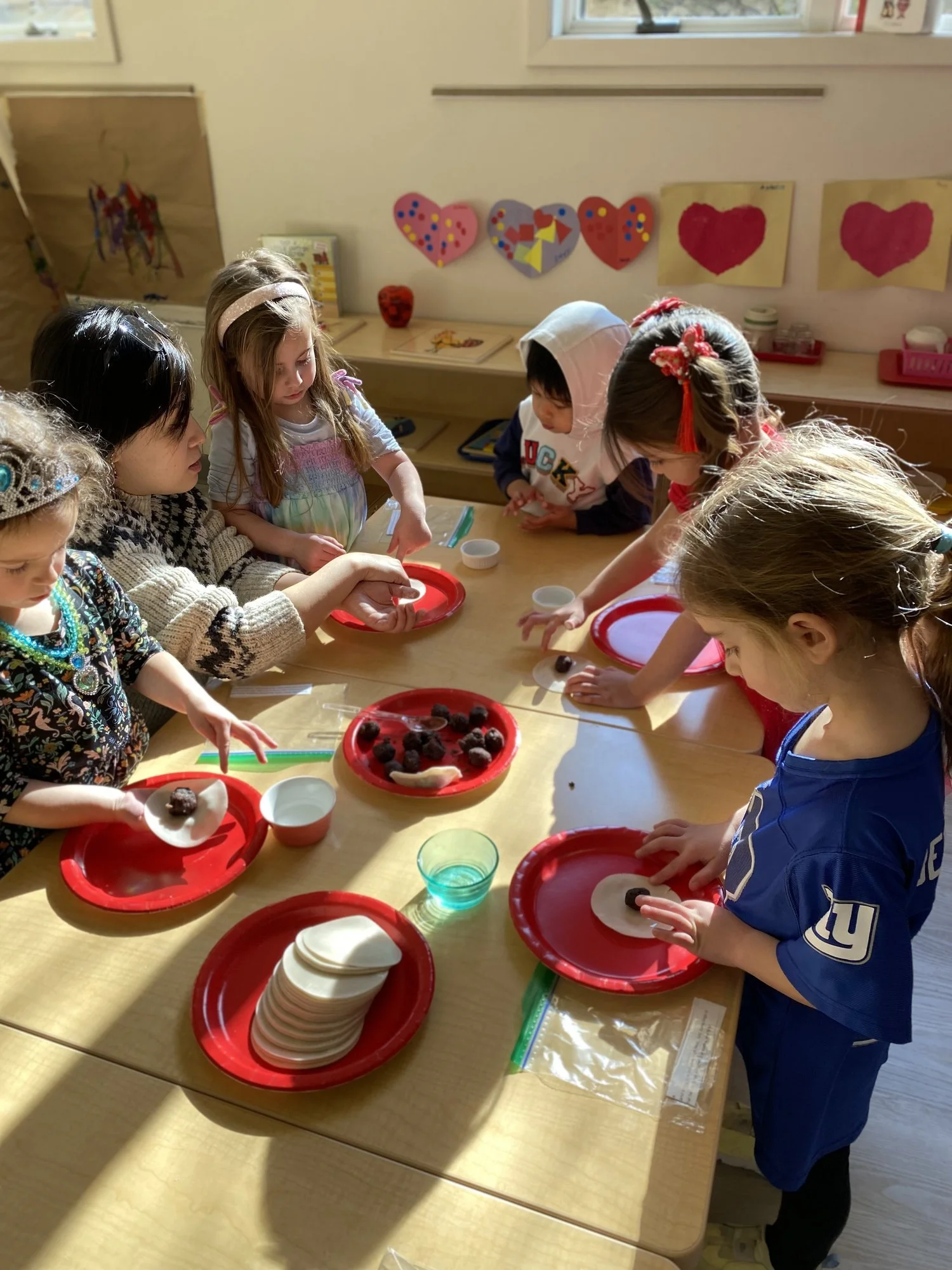 Children gathered around a table preparing food with dough and fillings on red plates, surrounded by heart-shaped decorations on the wall.