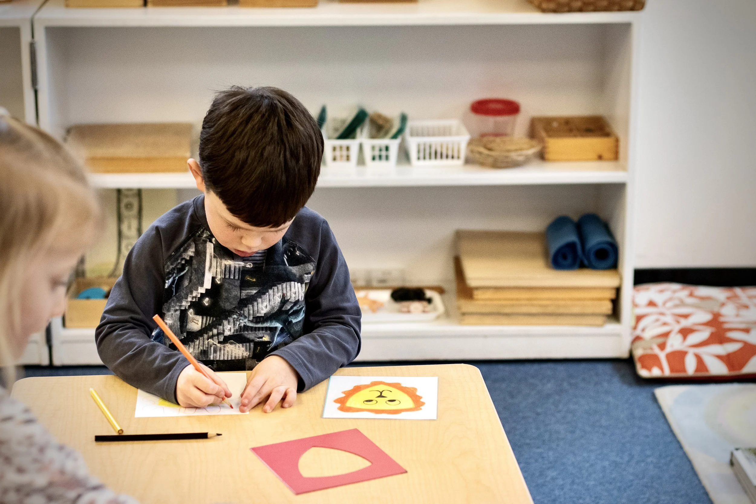 Children sitting at a table in a classroom, one drawing with a pencil on paper featuring a lion's face, shelves with educational materials in the background.