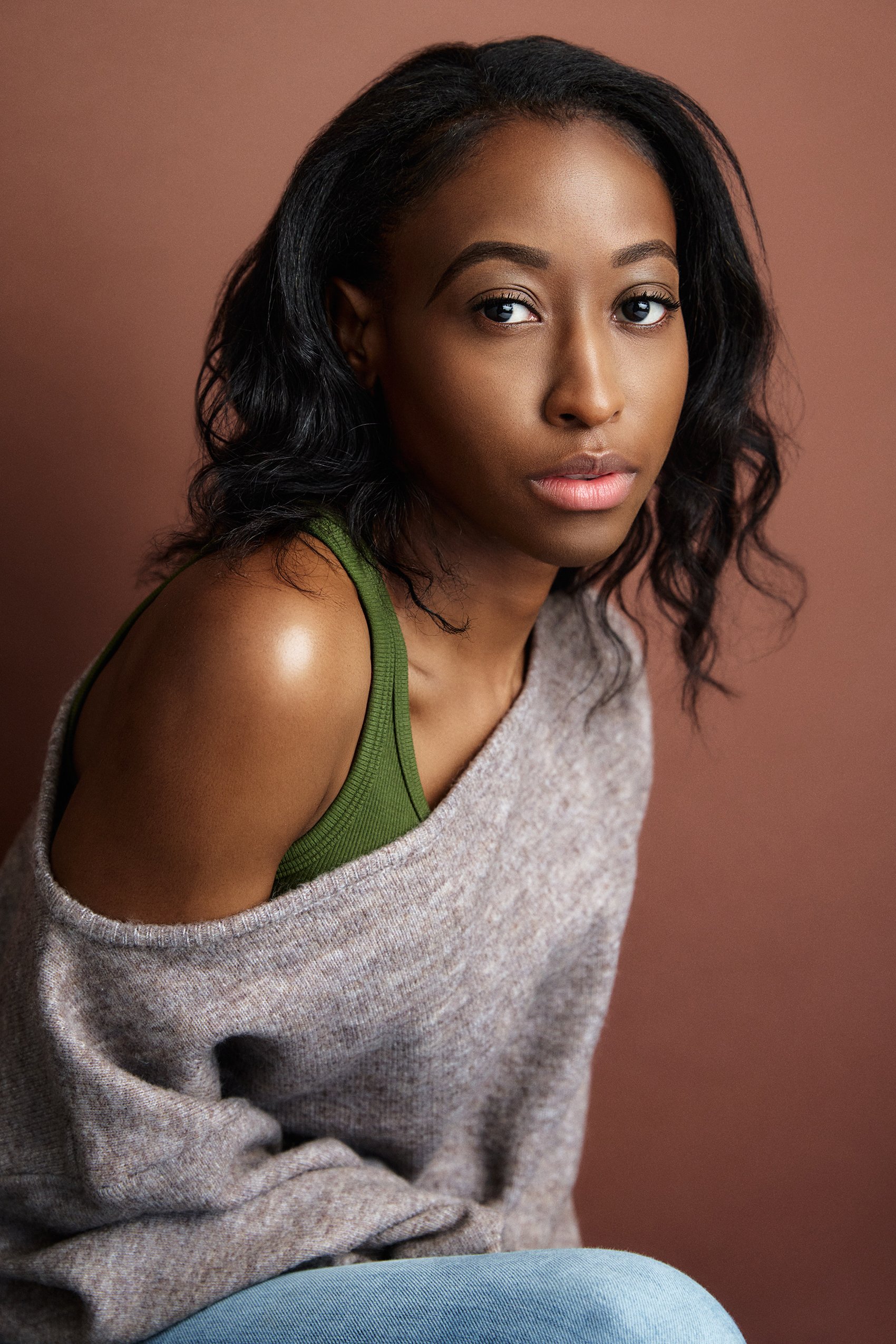 A young woman with dark wavy hair and brown skin, wearing a green tank top under a loose beige sweater, sits against a plain brown background, looking directly at the camera.